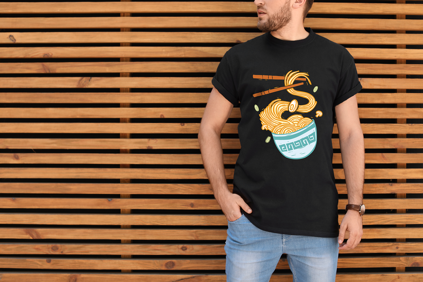 A man wearing a black T-shirt with a graphic of a bowl of ramen noodles held in chopsticks, standing in front of a wooden slatted wall.
