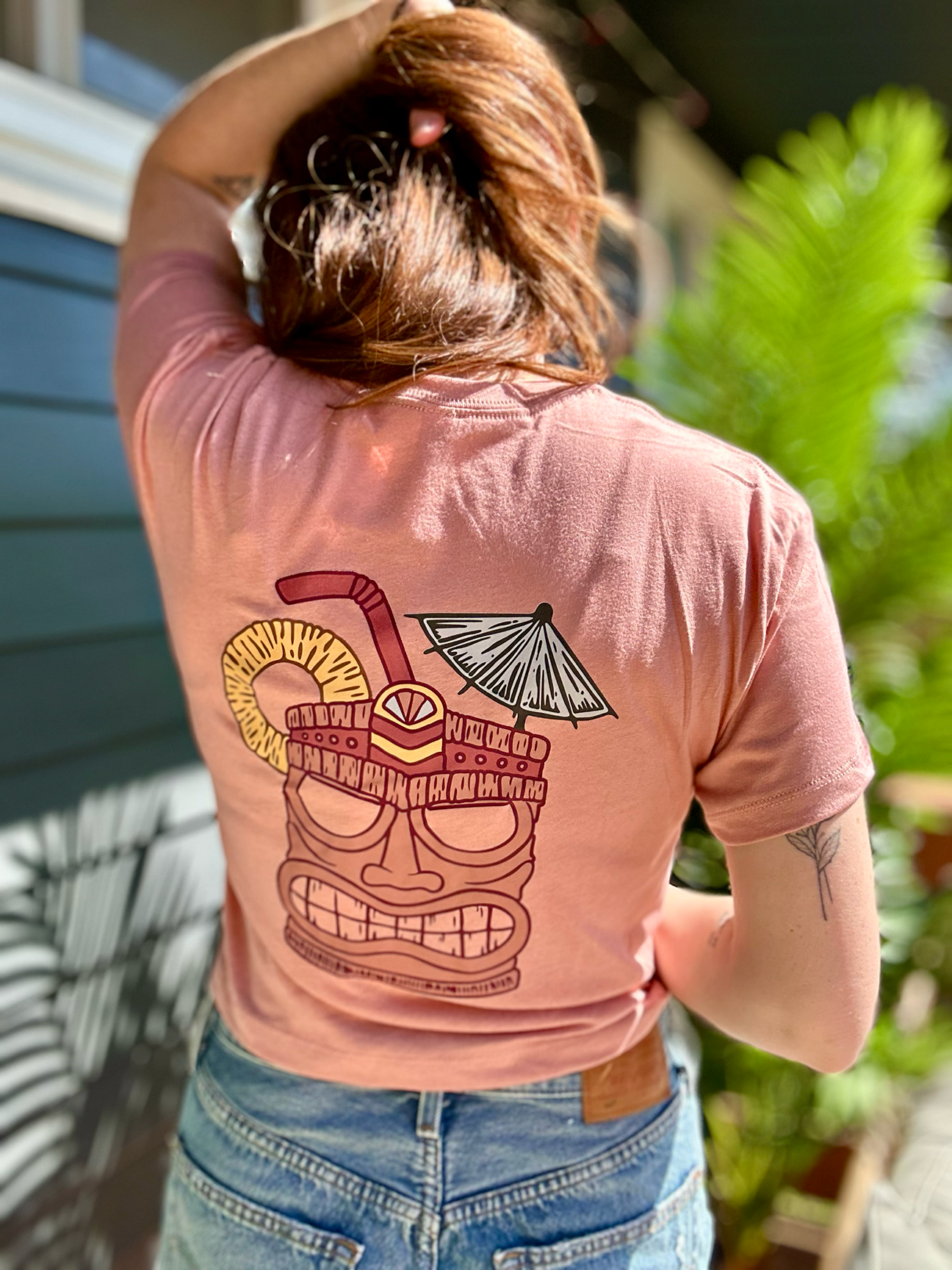 Back of a person wearing a peach-colored T-shirt with a tropical drink graphic on the back. The person has reddish hair and is outdoors near greenery and a house.