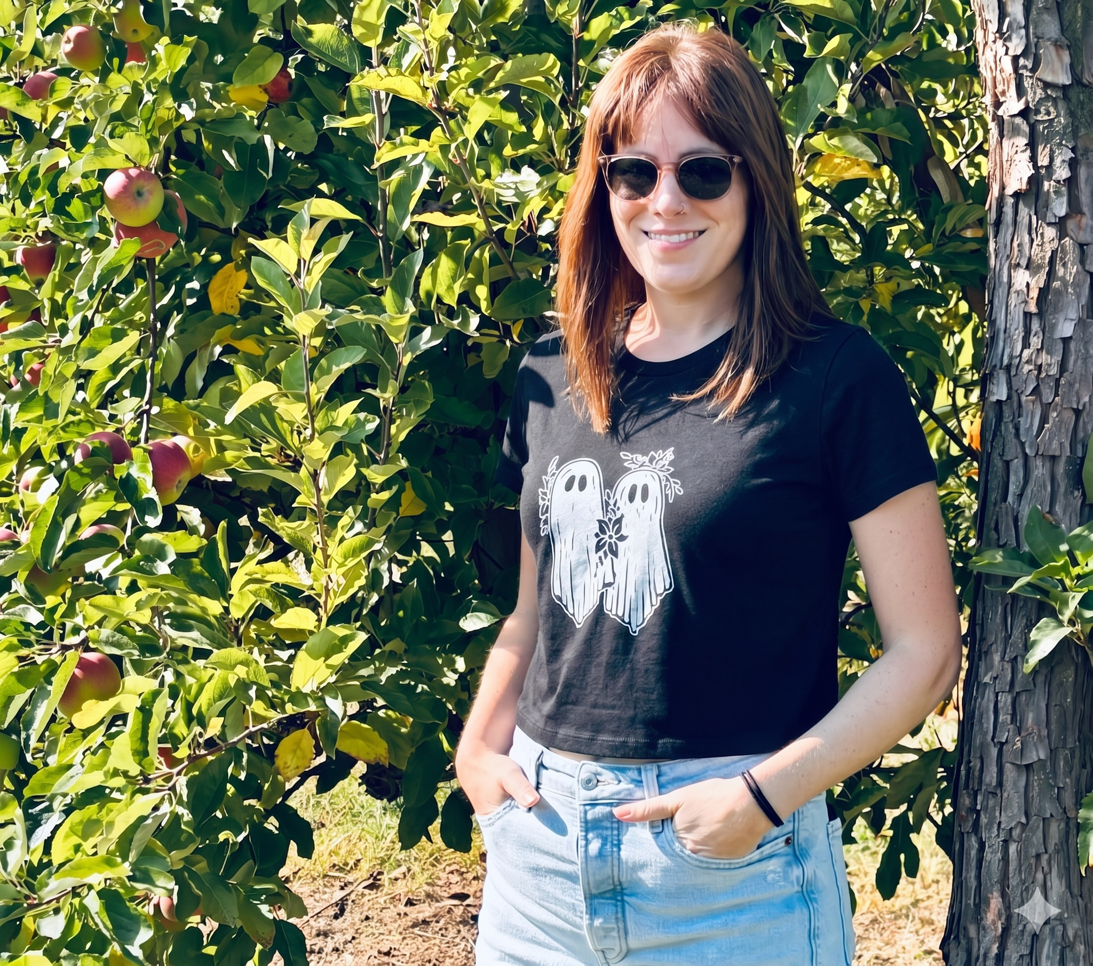 A young woman with brown hair and sunglasses standing outdoors next to an apple tree with green leaves and ripening apples, smiling at the camera.