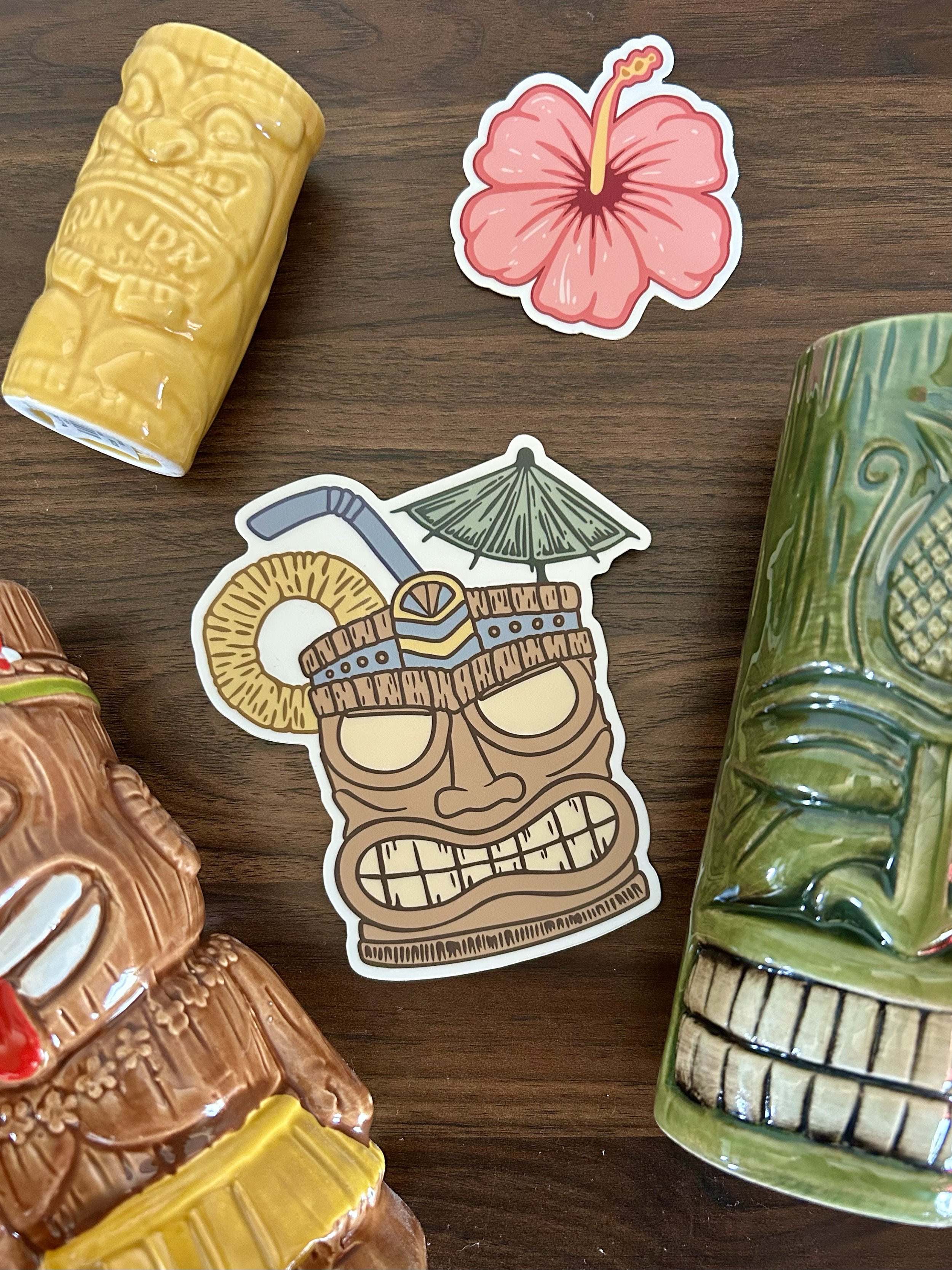 Colorful tiki-style stickers of tropical drinks, a hibiscus flower, and a ceremonial tiki figure on a wooden surface, surrounded by ceramic mugs.
