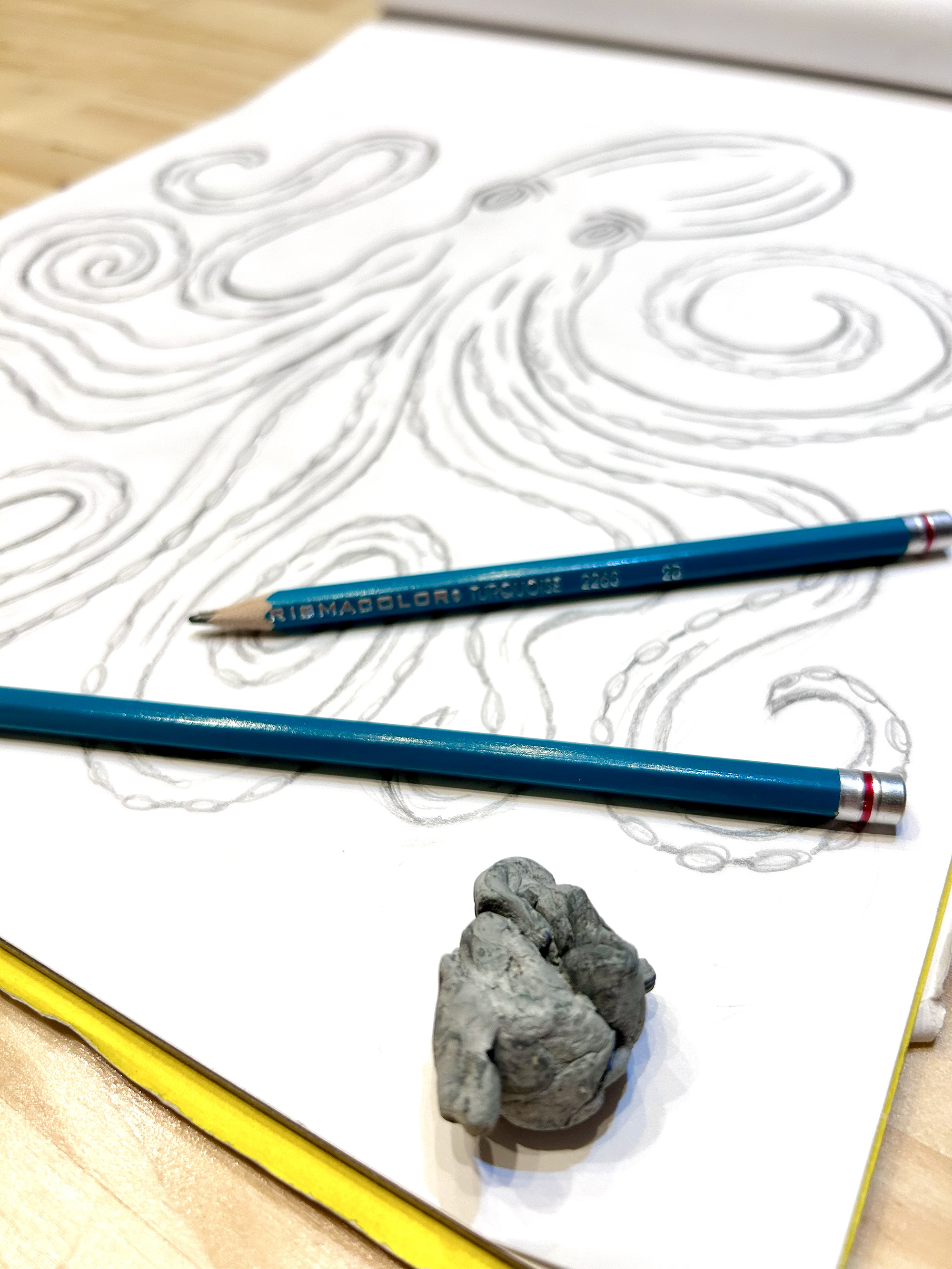 A sheet of paper with a swirling, flowing pattern drawn on it, two blue colored pencils lying across the paper, a small ball of gray clay, and a crumpled piece of gray clay.
