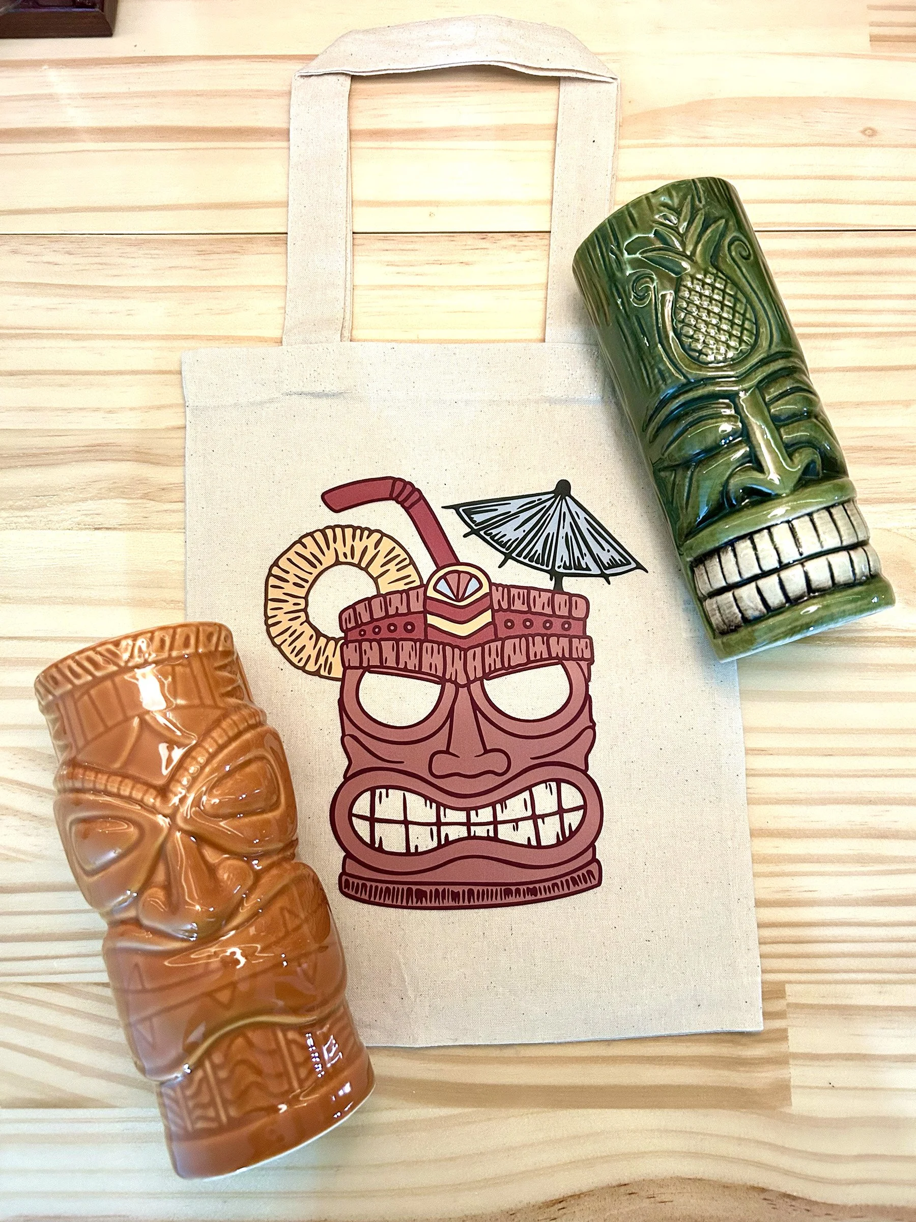 A decorative coconut-shaped mug and a ceramic tiki mug placed on a canvas tote bag with a Hawaiian drink illustration, all on a wooden surface.