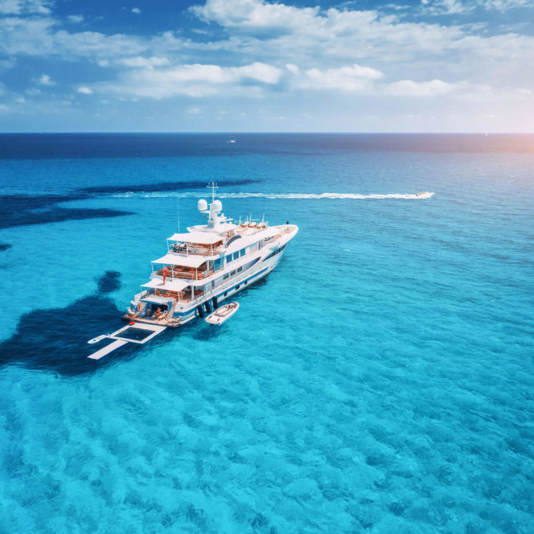 Aerial picture of a large yacht on crystal clear water under a cloudy blue sky.