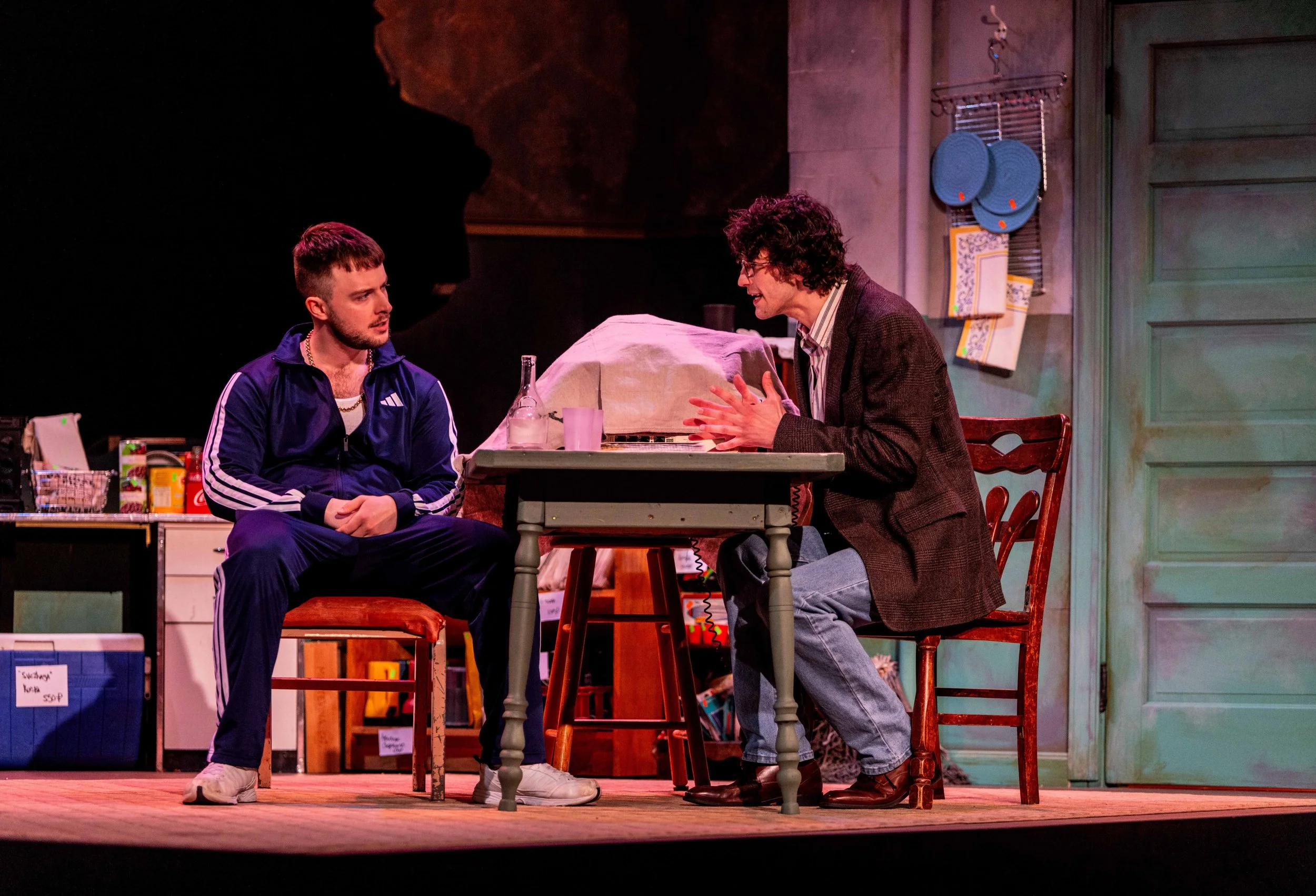 Two men sitting at a table on a stage, having a serious conversation. One is dressed in casual athletic wear, and the other in a blazer. The setting appears to be a theatrical play with a colorful background and cluttered scene.