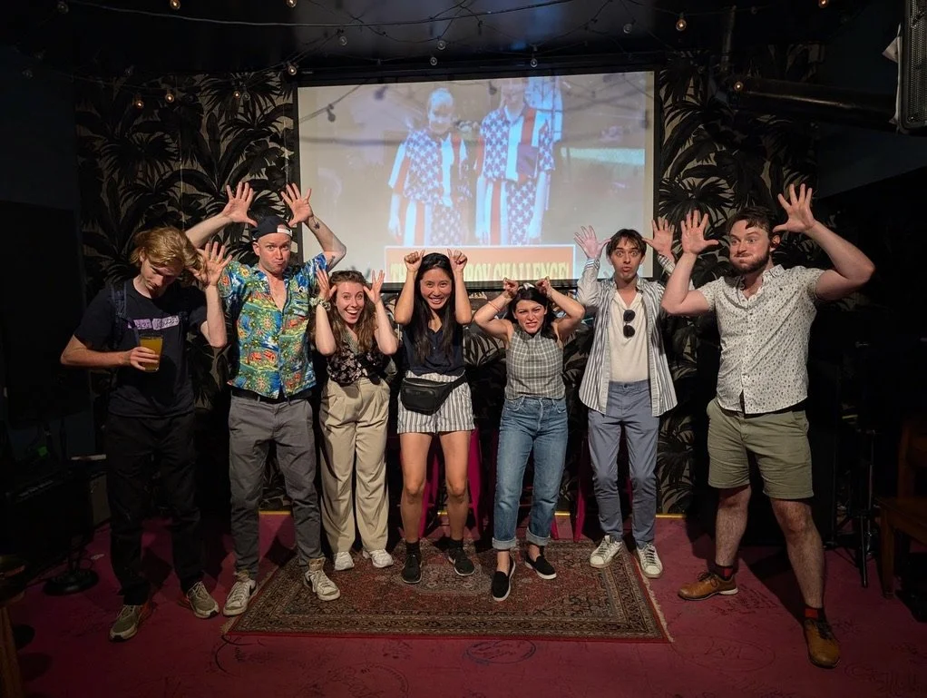 A group of seven young adults standing on a small stage in a dimly lit venue, posing with hands raised. The group is celebrating an improv performance.