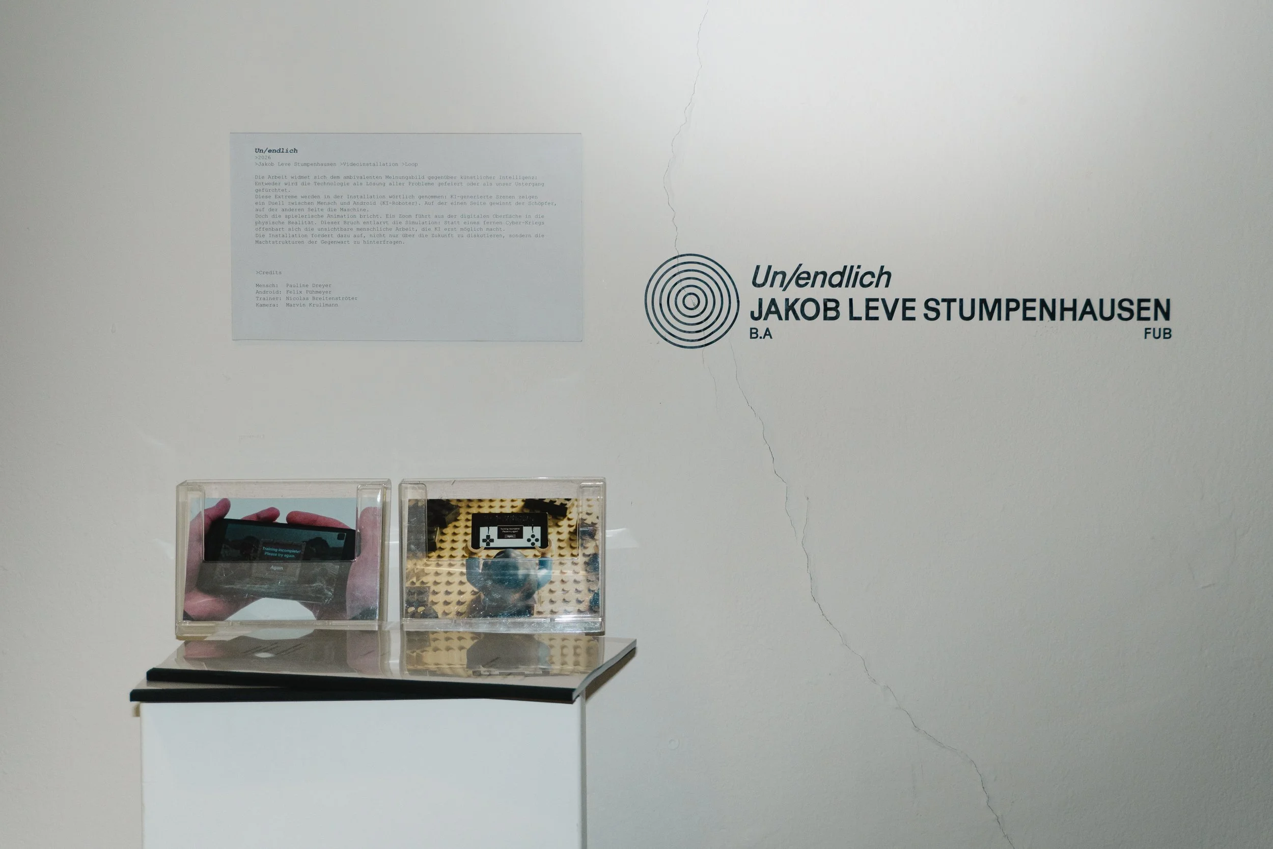 Ausstellungstafel mit schwarzem Spiral-Design und Text, darunter einen kleinen Bildschirm, der eine Hand mit Smartphone zeigt. Es gibt auch eine kleine Spielfigur in einer transparenten Box.