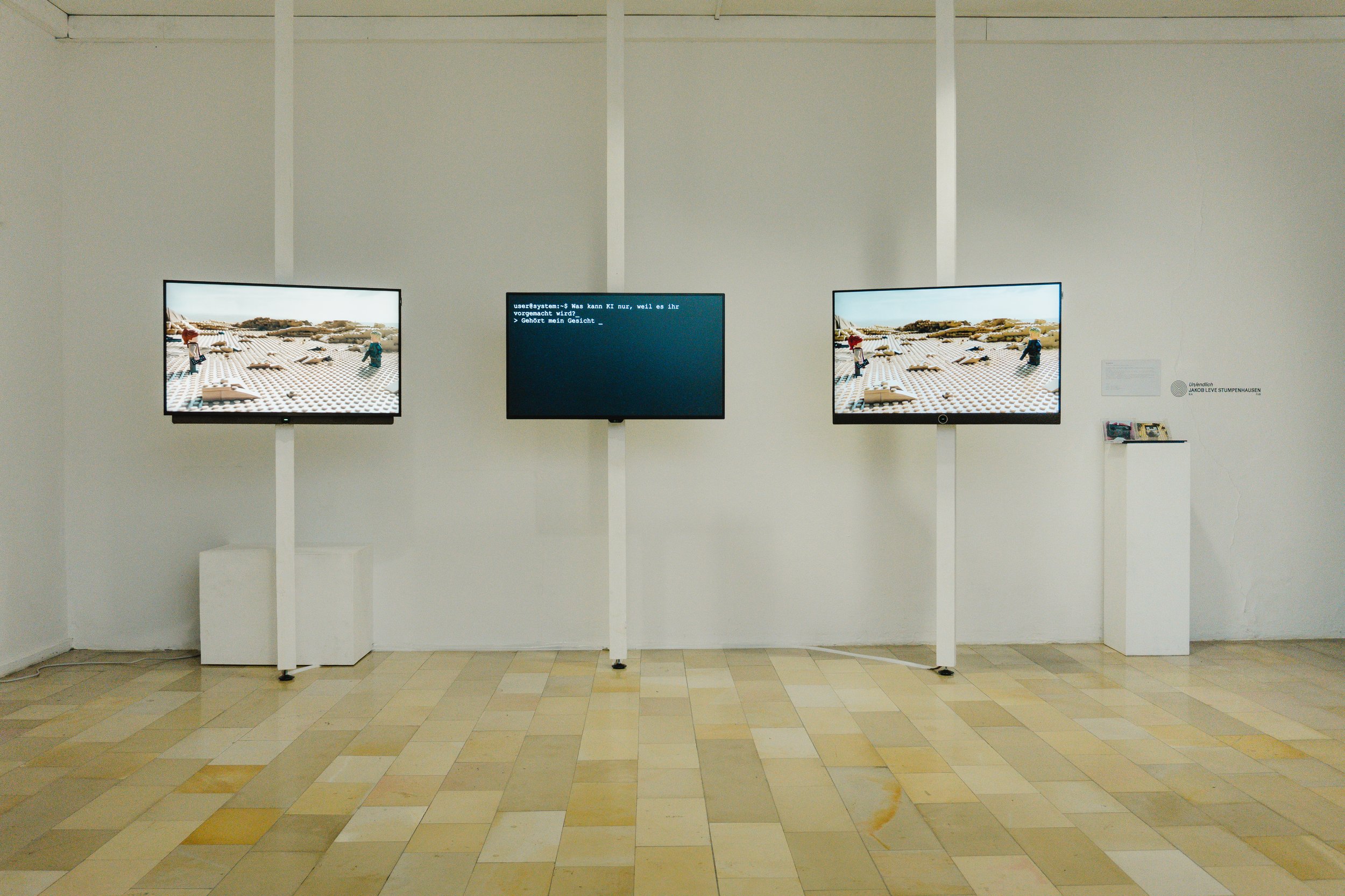 Drei Bildschirme in einer Kunstgalerie, die ein Lego-Sandkasten mit Lego-Figuren zeigen, mit einem Bildschirm in der Mitte, der einen Computercode darstellt.