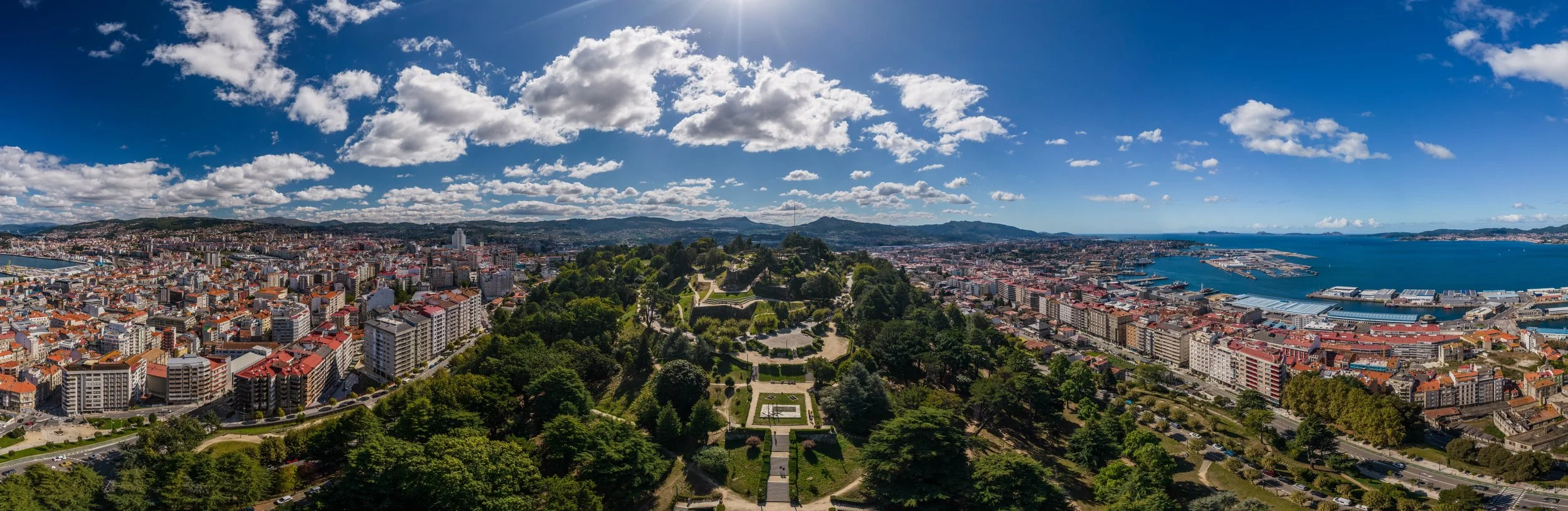 Vista panorámica de una ciudad costera como Vigo en España con un parque grande y áreas verdes en el centro, edificios residenciales y comerciales, y un puerto con barcos al lado del mar bajo un cielo parcialmente nublado.