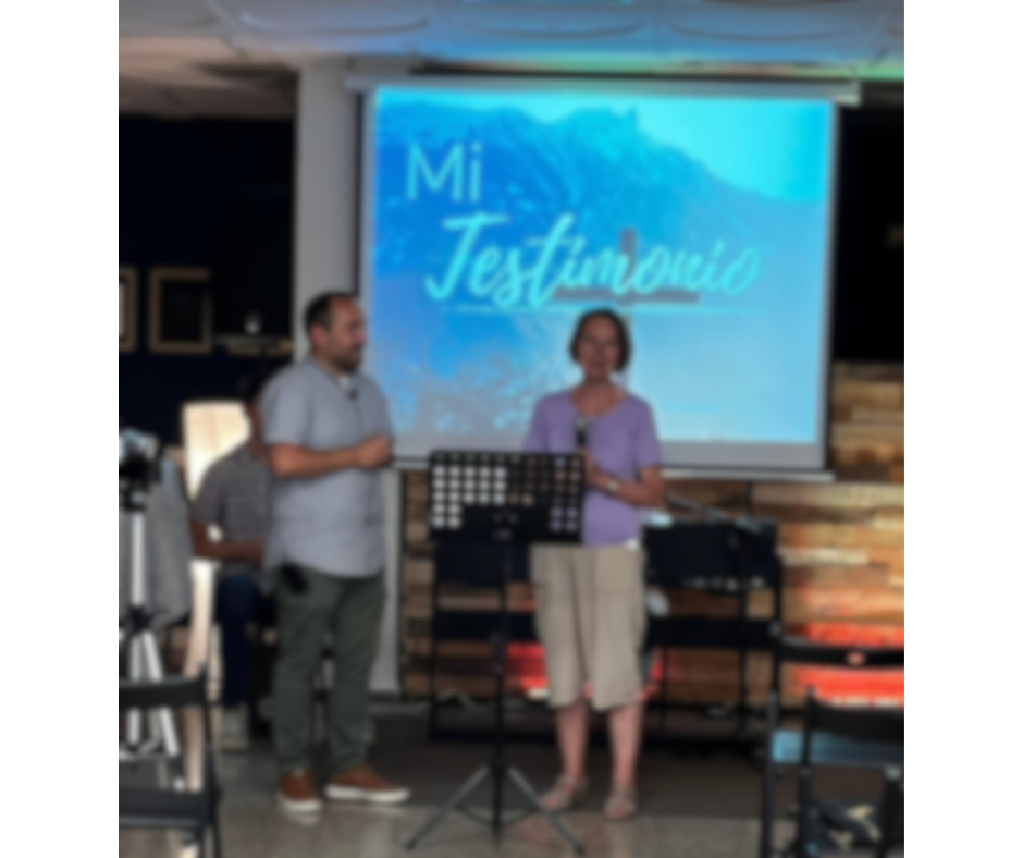 Dos personas de Vigo comunidad Cristiana en un escenario participando en una ceremonia de testimonios, con un proyector que dice 'Mi Testimonio' en el fondo.