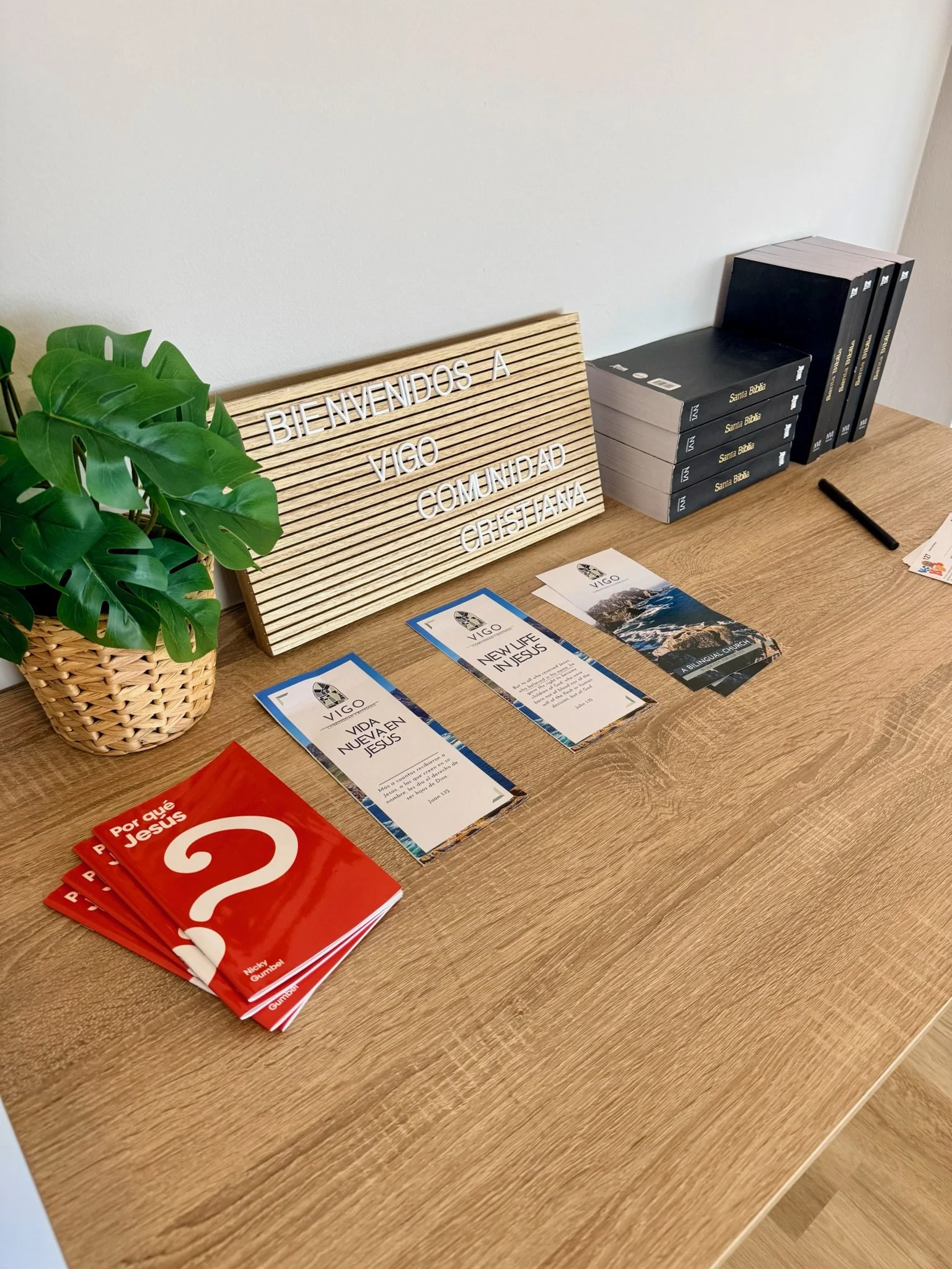 A wooden table with a small potted green plant, a letter board with the message 'Bienvenido a Vigo Comunidad Cristiana,' several brochures about Vigo, and a stack of red booklets titled 'Por qué Jesús'.