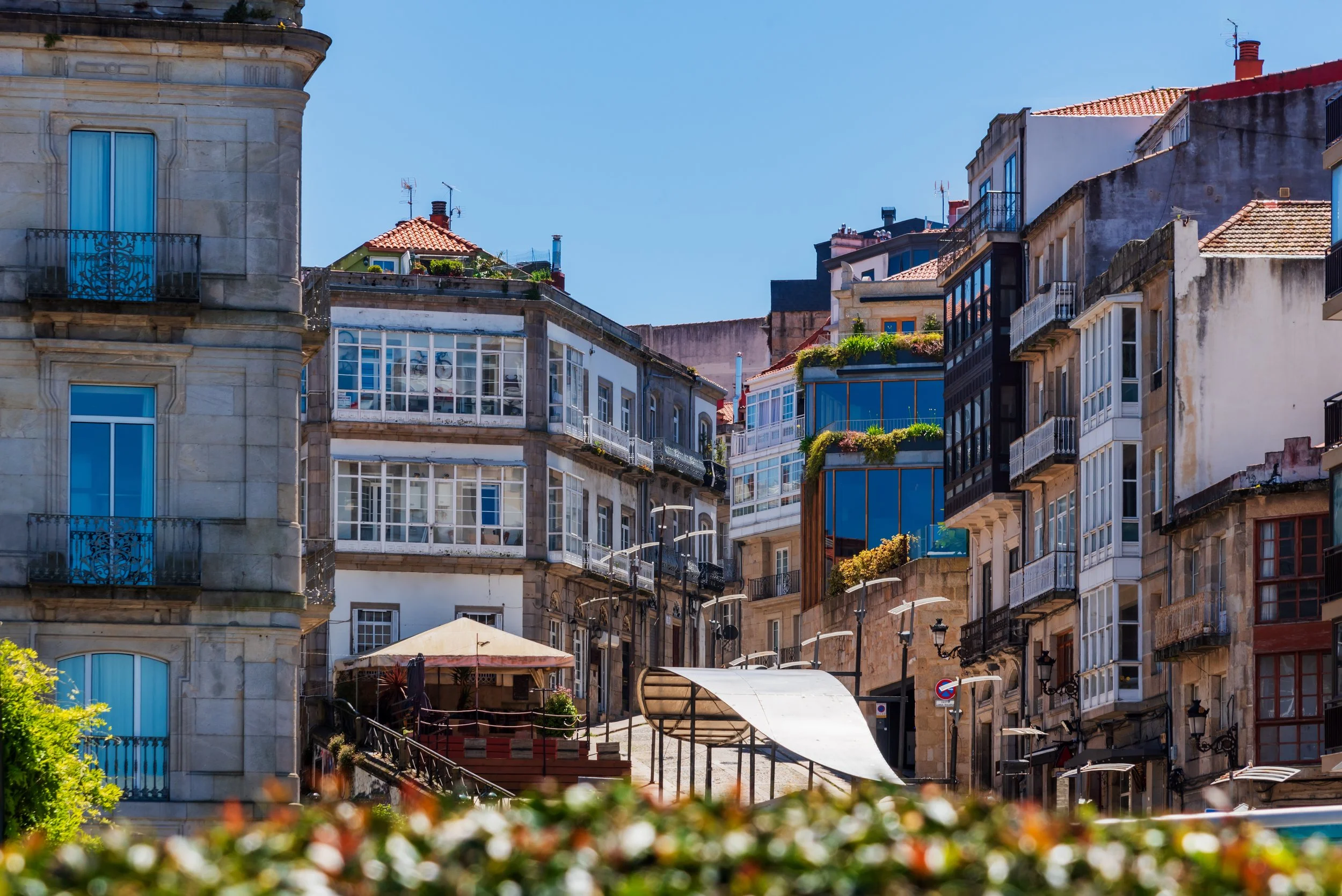 Vista de edificios residenciales en una calle en una ciudad de Galicia con algunos apartamentos con balcones y terrazas, y un cielo despejado.