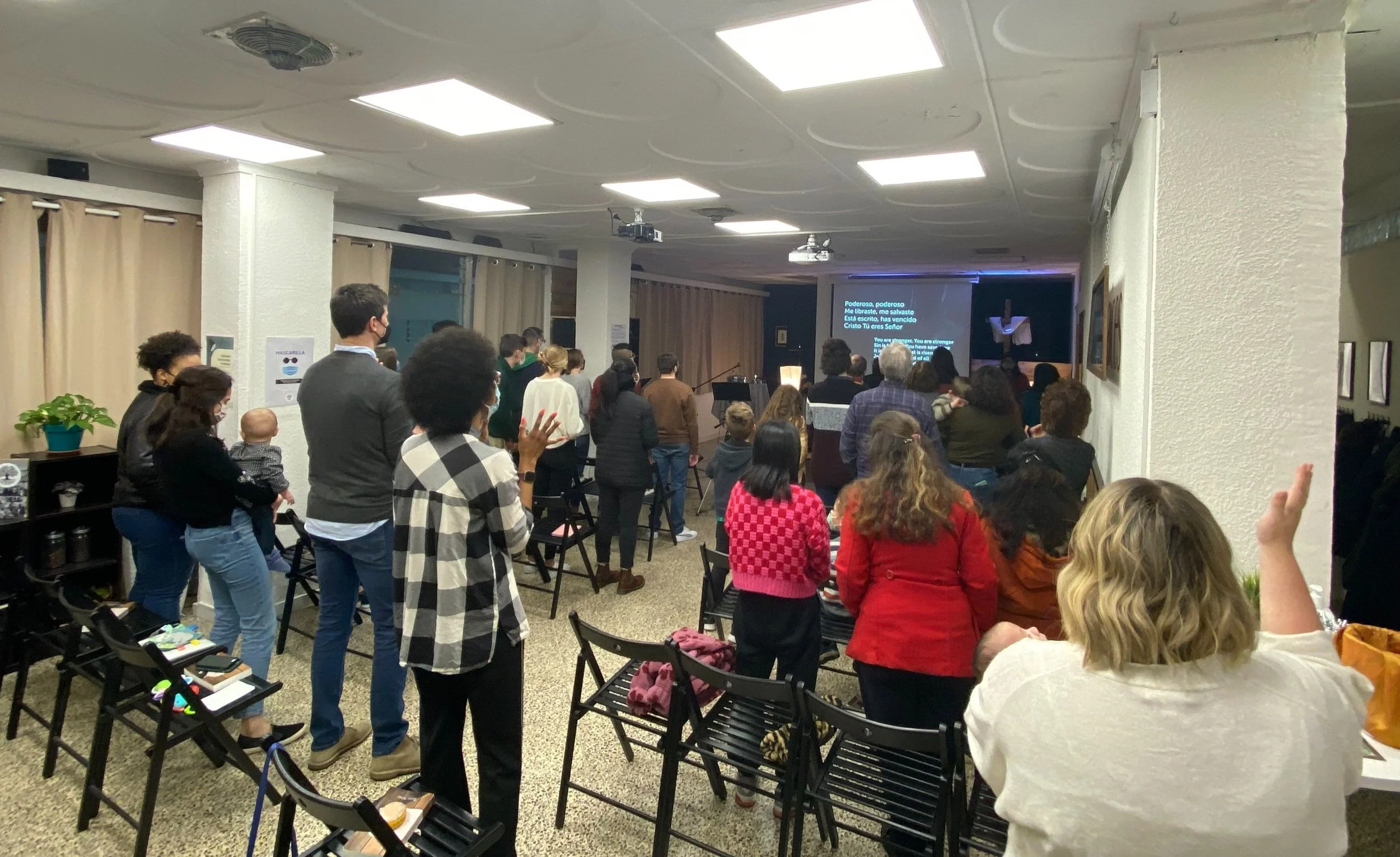 Personas congregadas en un salón, algunas de ellas de pie y otras sentadas, participando en un evento religioso con un altar y una cruz, mientras en la pantalla se proyectan letras en español e inglés de una canción o himno religioso.