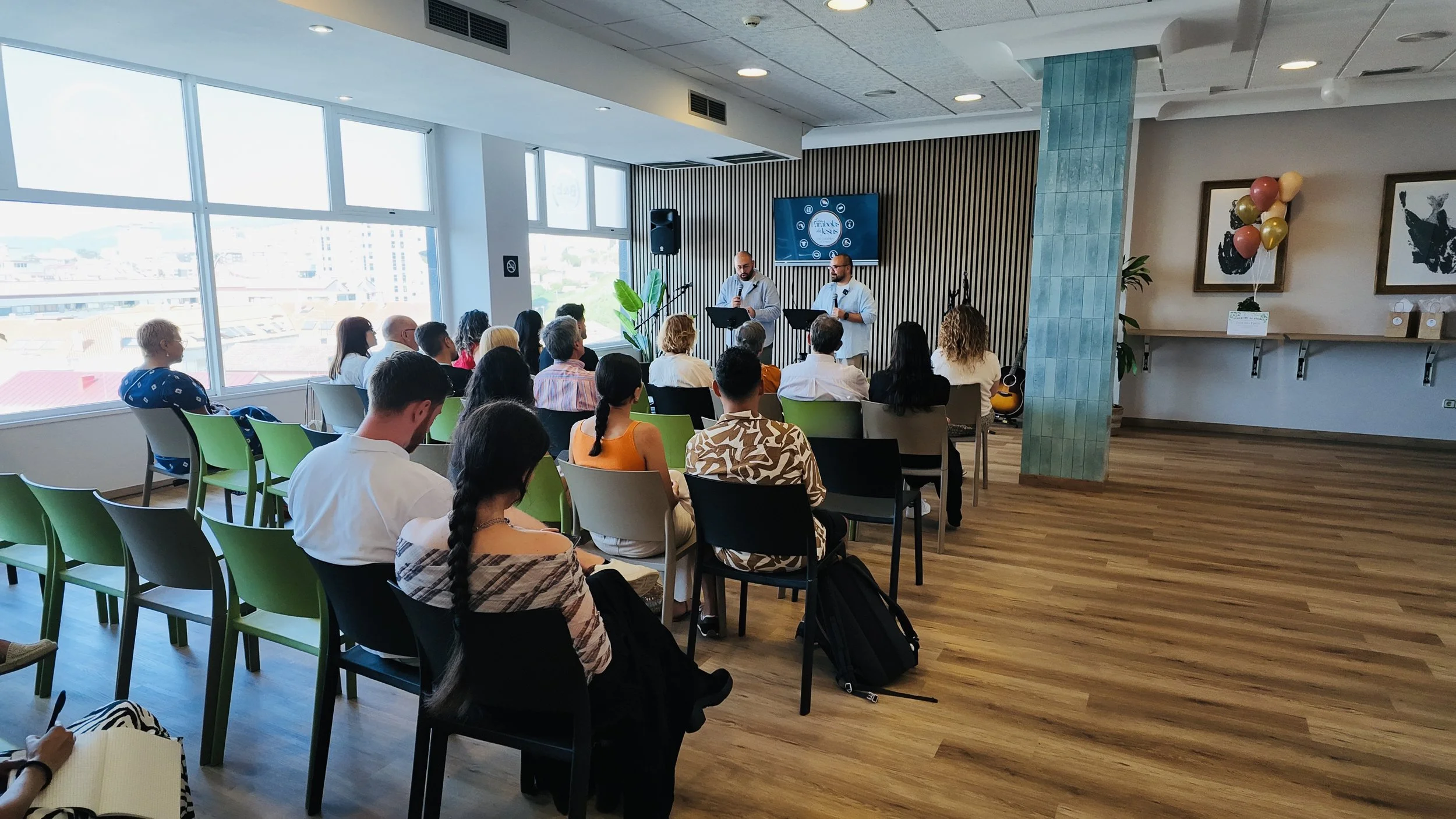 People attending a presentation in a bright, modern room with large windows, seated in green and black chairs, listening to two speakers standing in front of a digital screen Vigo comunidad cristiana Spain.