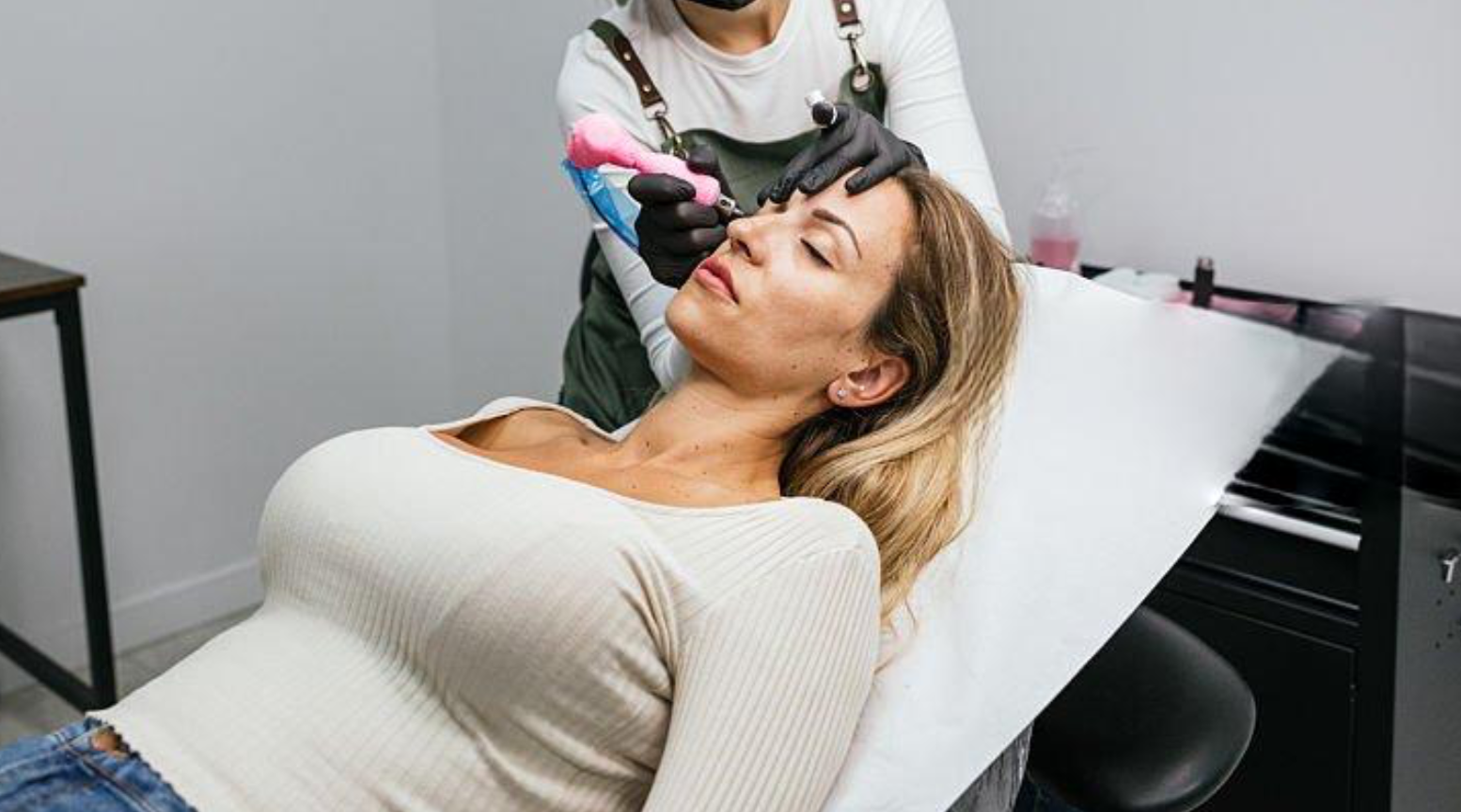 A woman lying on a chair with her eyes closed while a tattoo artist creates a tattoo on her forehead.