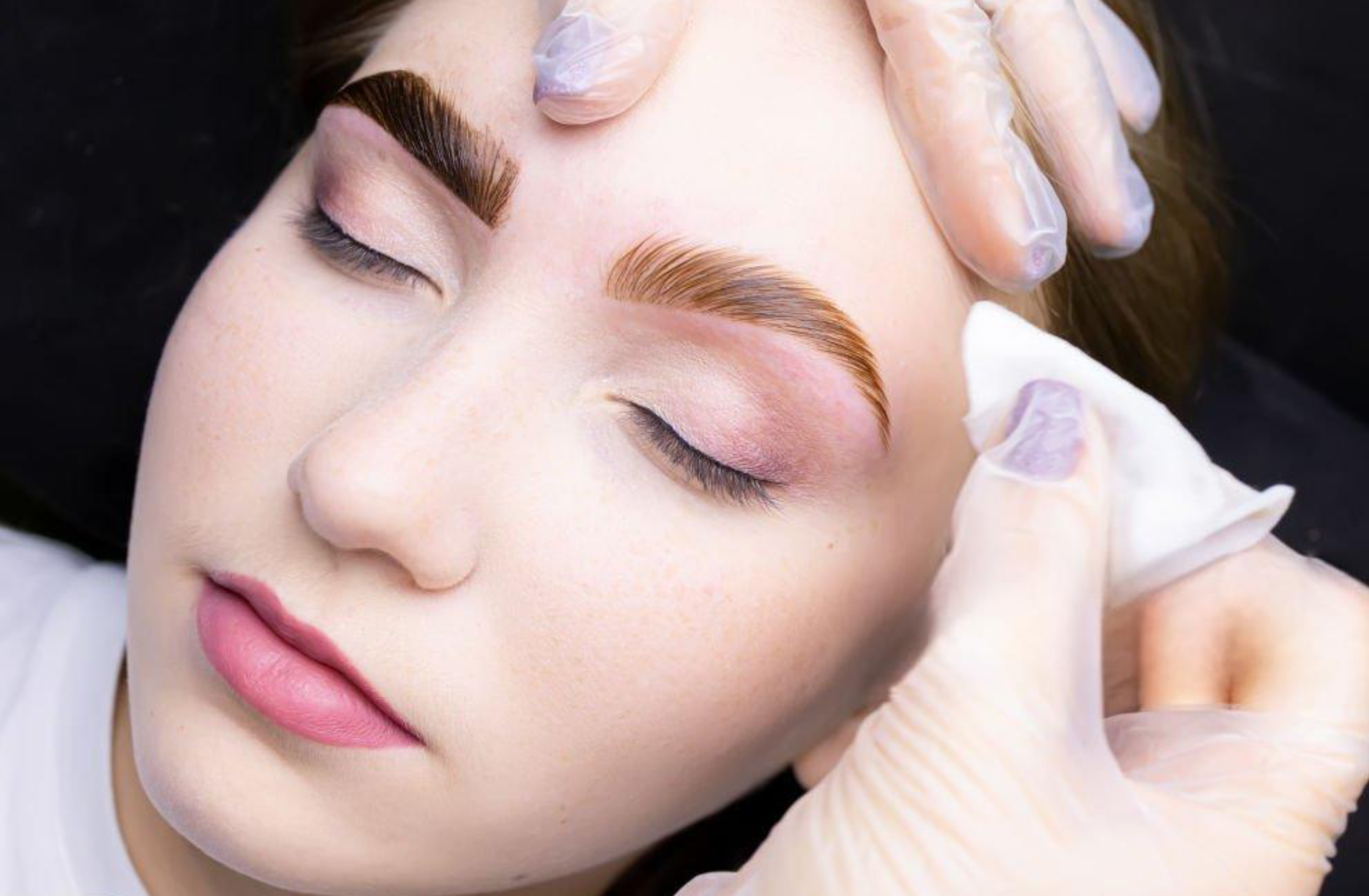 A woman with closed eyes receiving a facial treatment, with a professional wearing gloves gently touching her forehead.