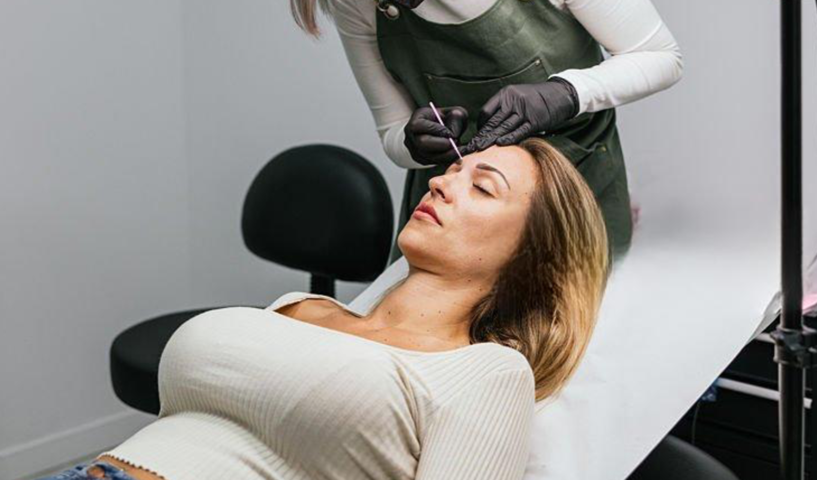 A woman had her eyes closed during a permanent makeup procedure in a tattoo studio. The tattoo artist is wearing gloves and is using a tattoo needle.
