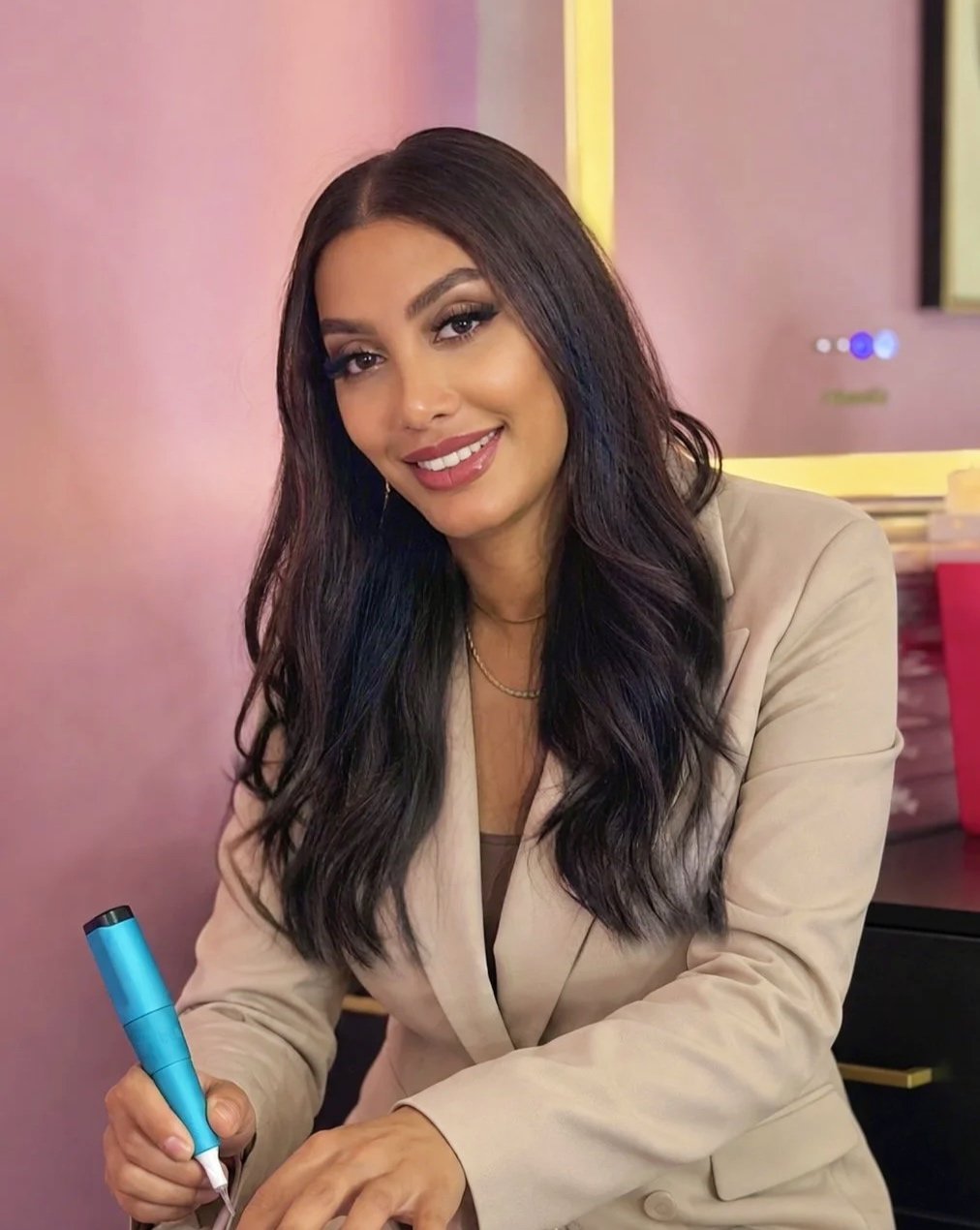 A woman with long dark hair and makeup, smiling and holding a tattoo machine, sitting at a table in a colorful room.