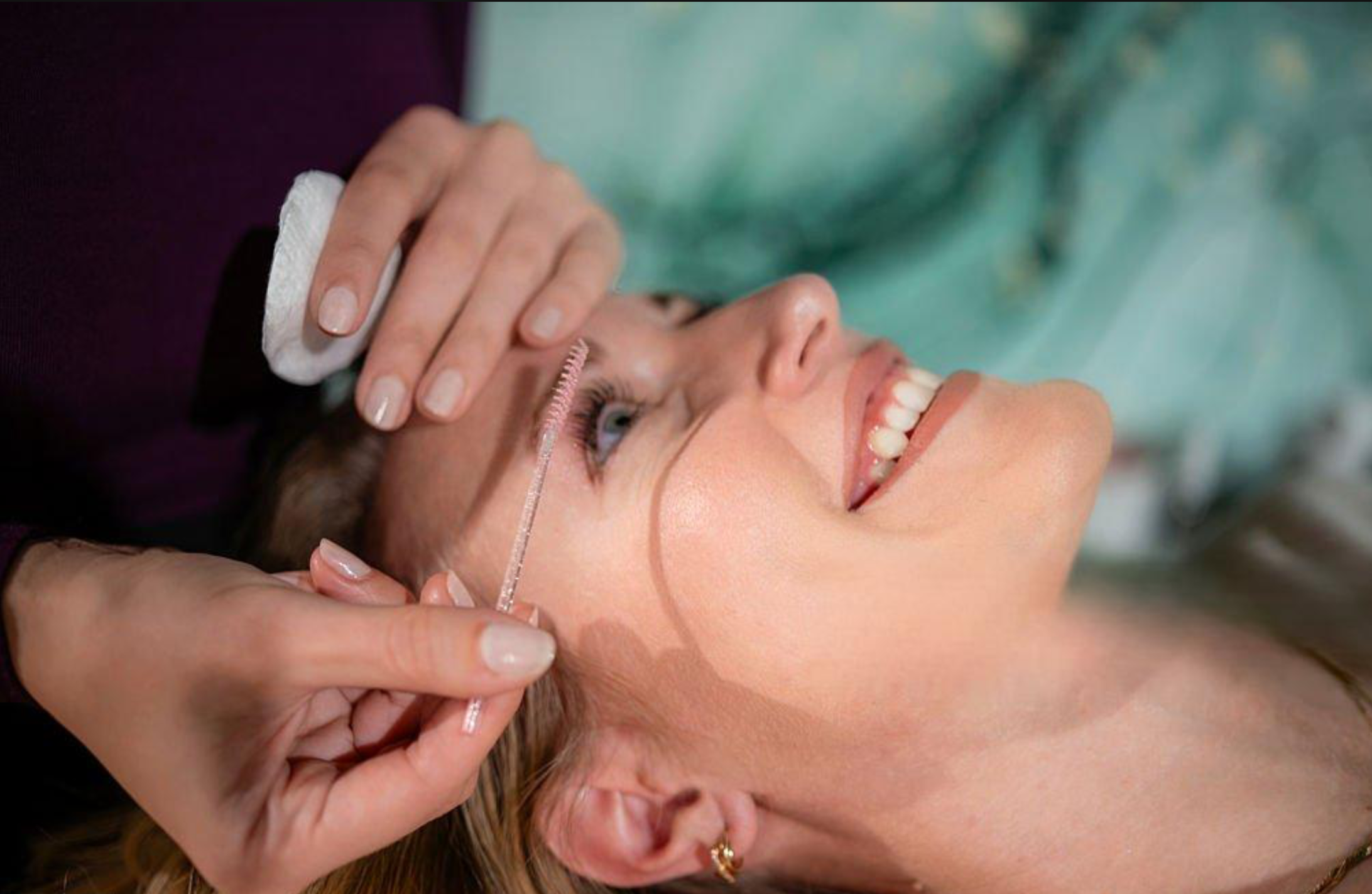 A woman lying in a medical setting, smiling, with an artist performing an eyebrow lamination procedure.