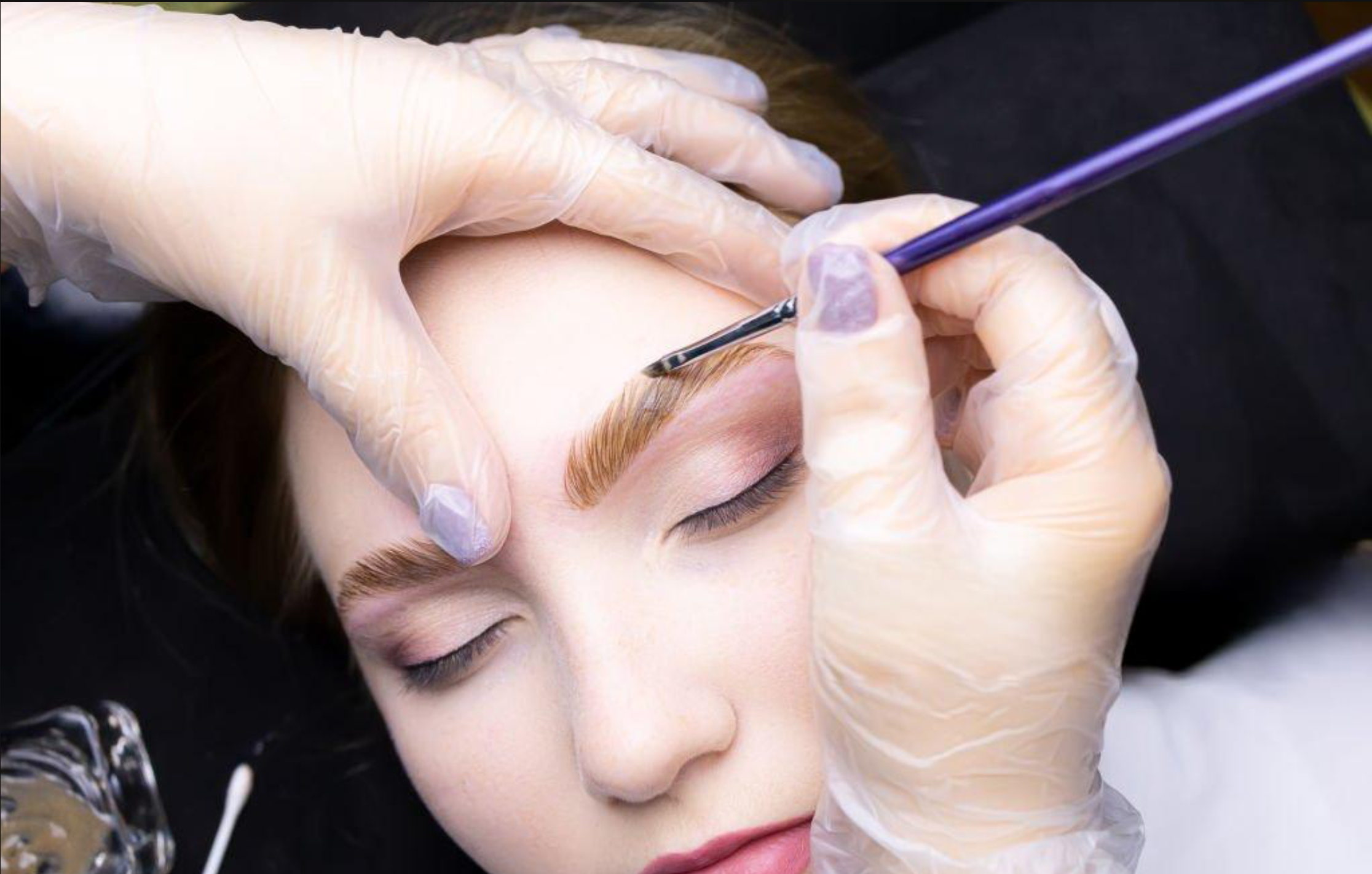A person with closed eyes receives a cosmetic or medical procedure on their brow from a practitioner wearing gloves, using a small tool.