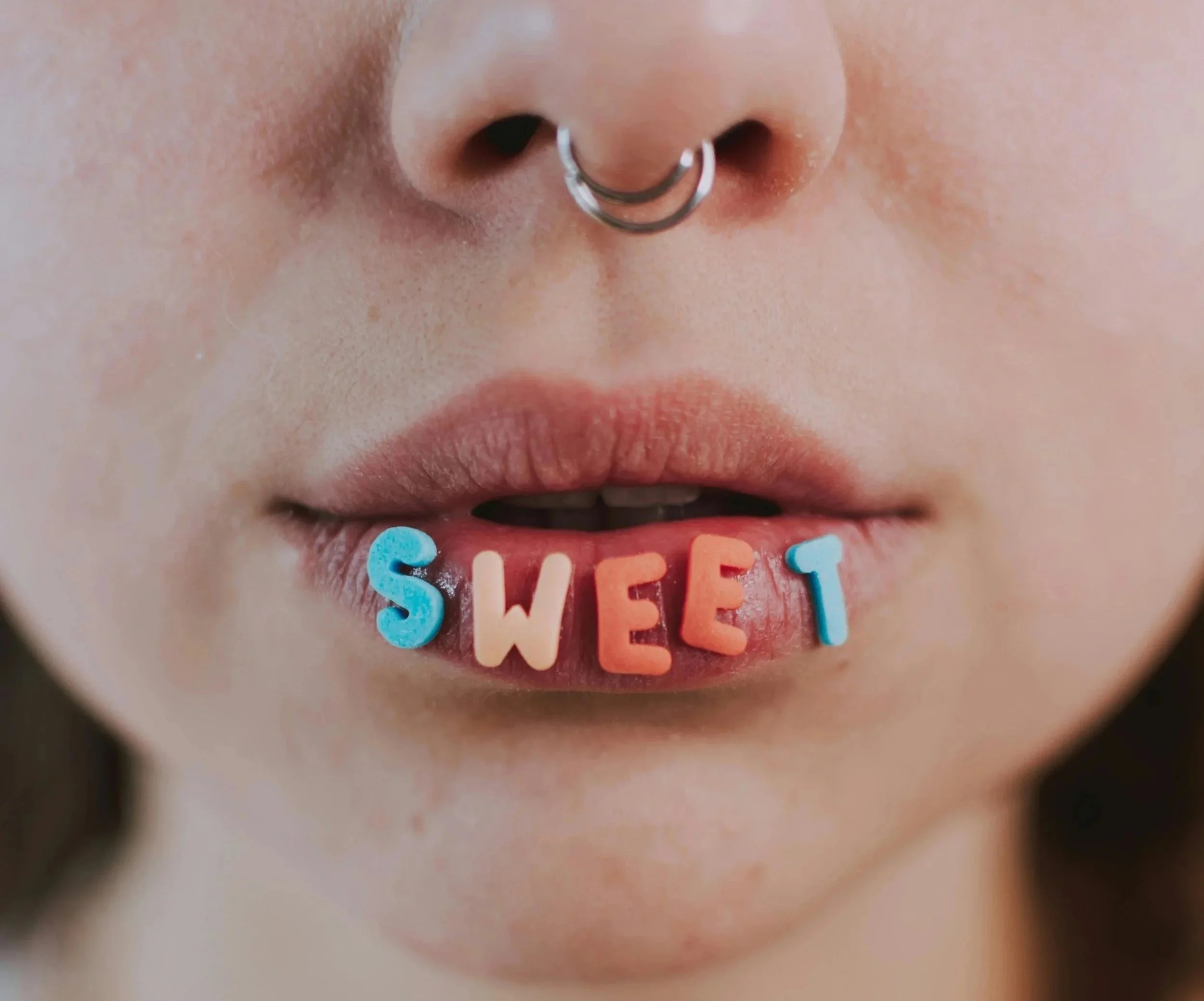 Close-up of a person's mouth with colorful letter-shaped candies spelling out the word 'SWEET' and a nose piercing.