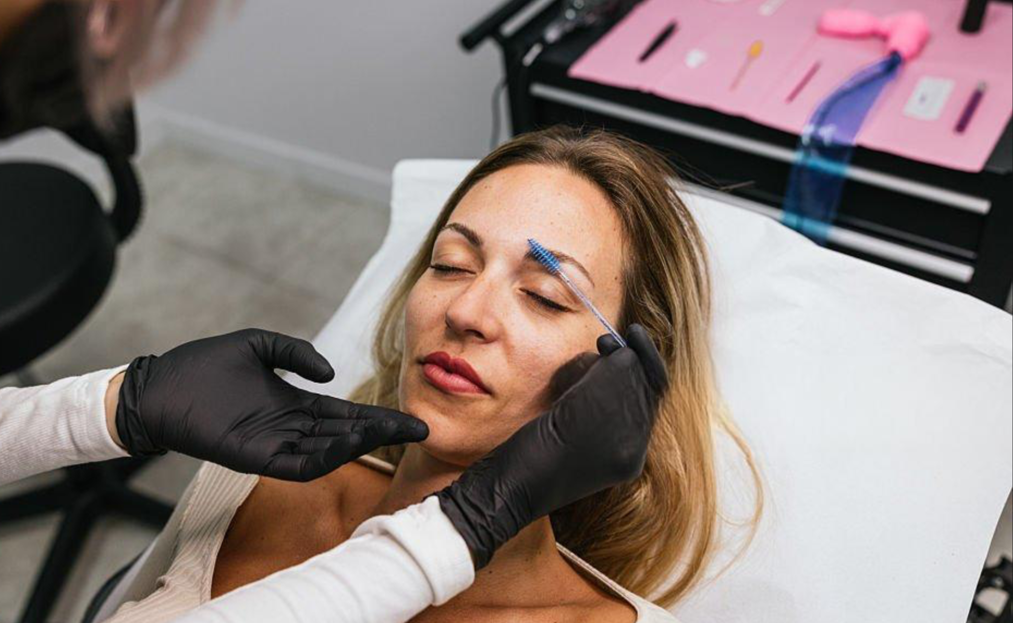 A woman with blonde hair and closed eyes receiving an eyebrow tattoo or microblading procedure by a technician wearing black gloves, with various tools and supplies on a pink mat nearby.