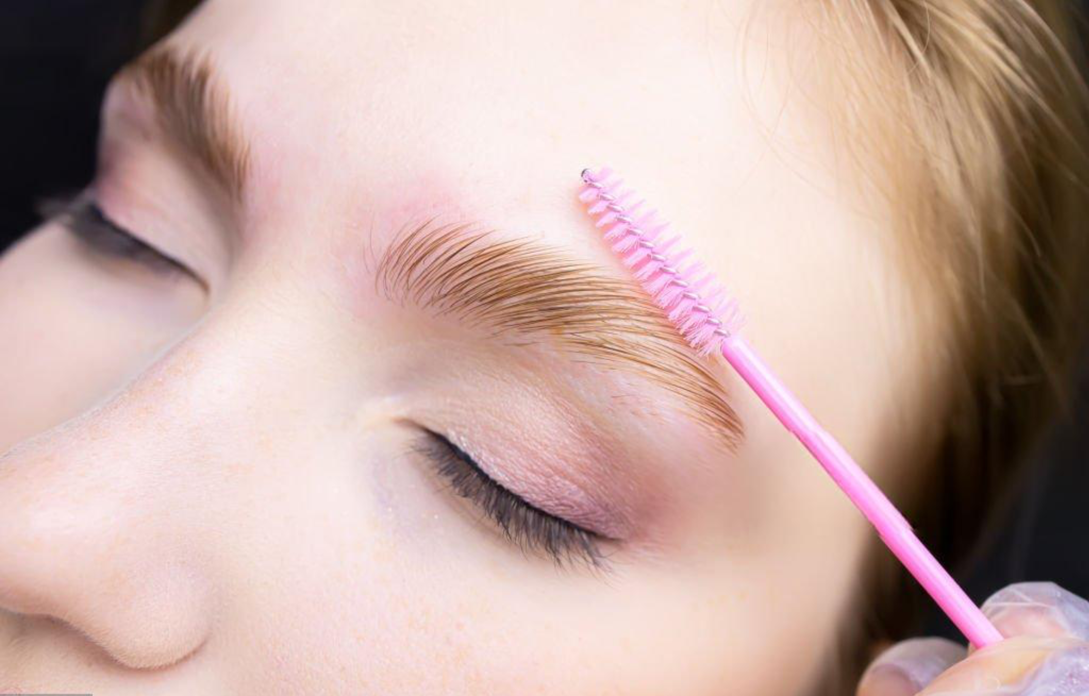 Close-up of a person with blonde hair getting eyelash extensions, with a pink cleaning brush nearby.