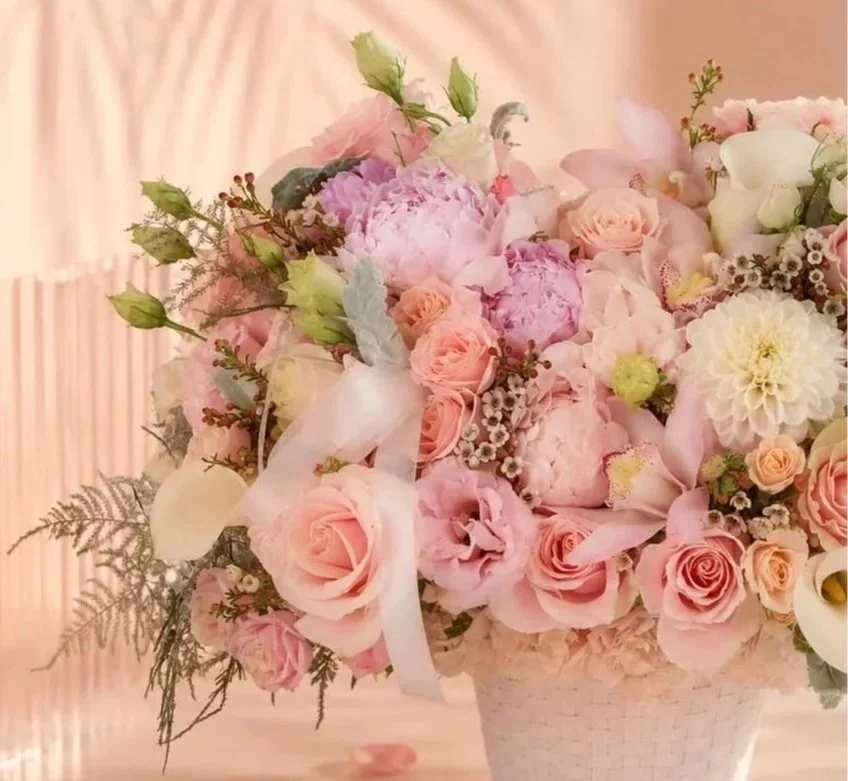 Close-up of a pink and white floral arrangement in a white basket, featuring roses, peonies, calla lilies, and other blooms with greenery.