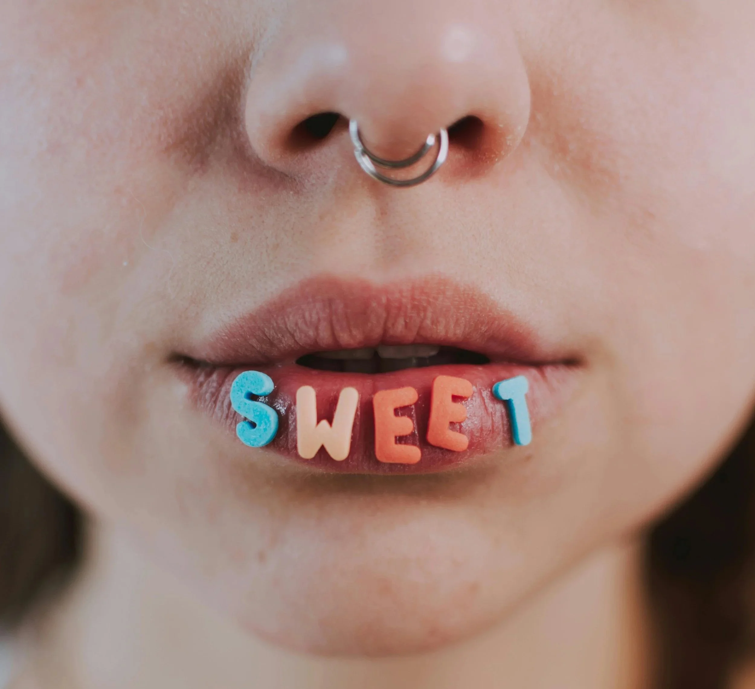 Close-up of a person's nose, lips, some colored letters spelling 'SWEET' hanging from the mouth, and a septum piercing.