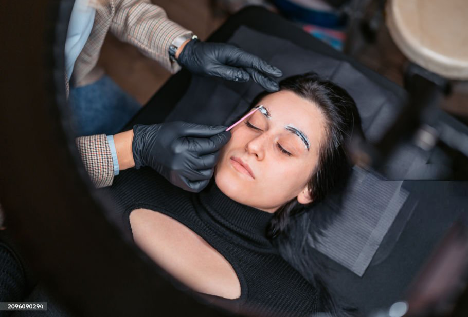 A woman with dark hair lying down, receiving an eyelash extension or makeup application from a professional wearing black gloves.