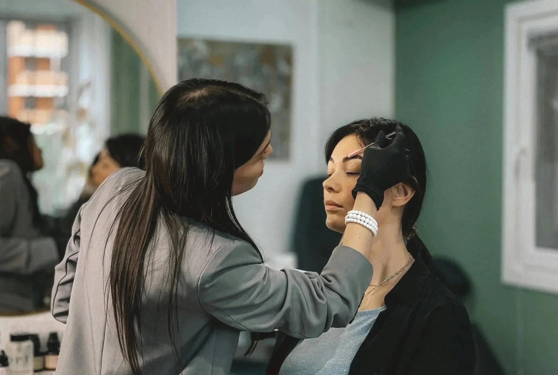 A woman with long dark hair applying makeup to another woman's face with a small brush in a green-walled room.