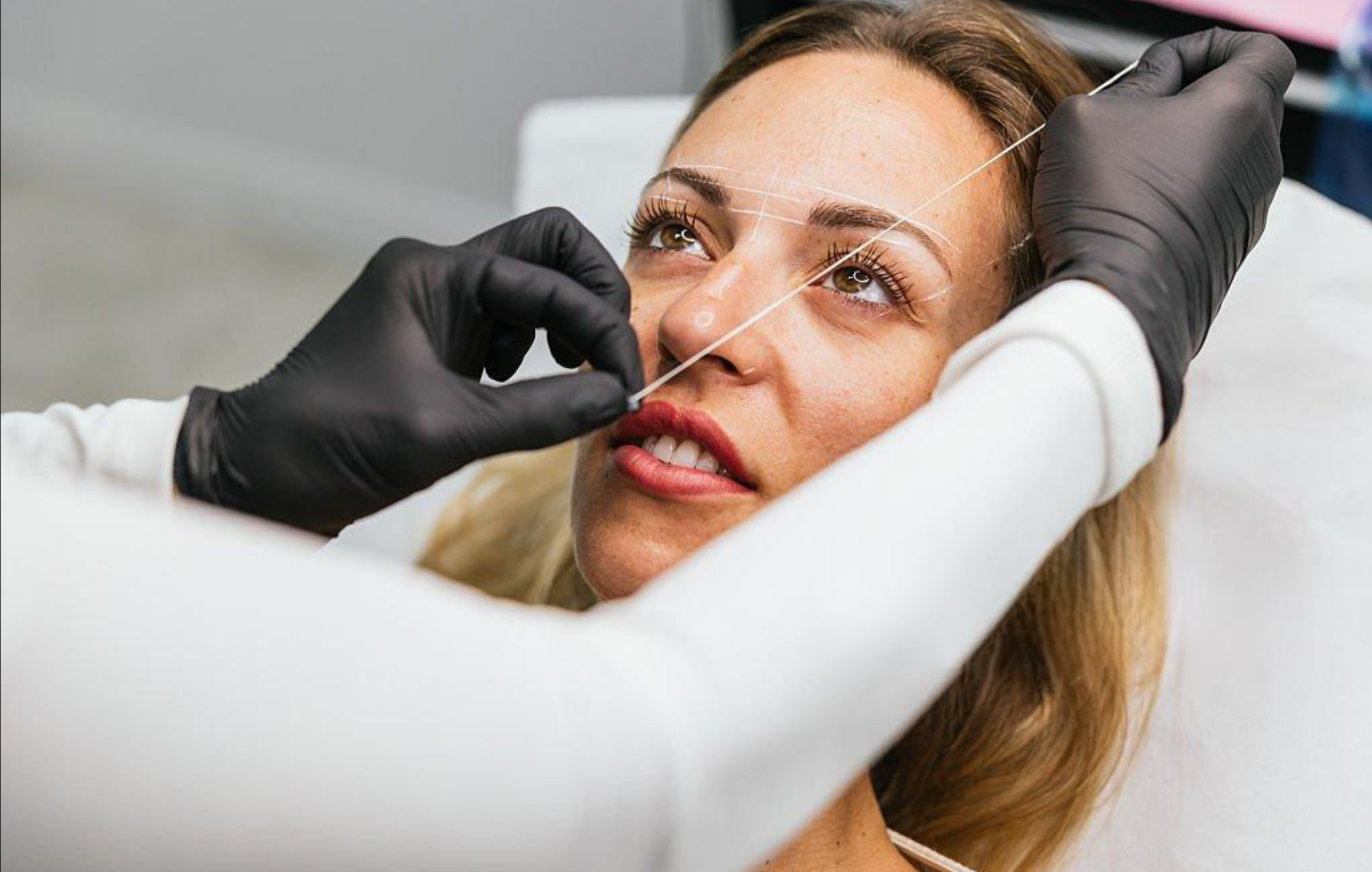 A woman with long brown hair lies down as a professional performs a cosmetic procedure on her face, using a needle and measuring lines for precision.
