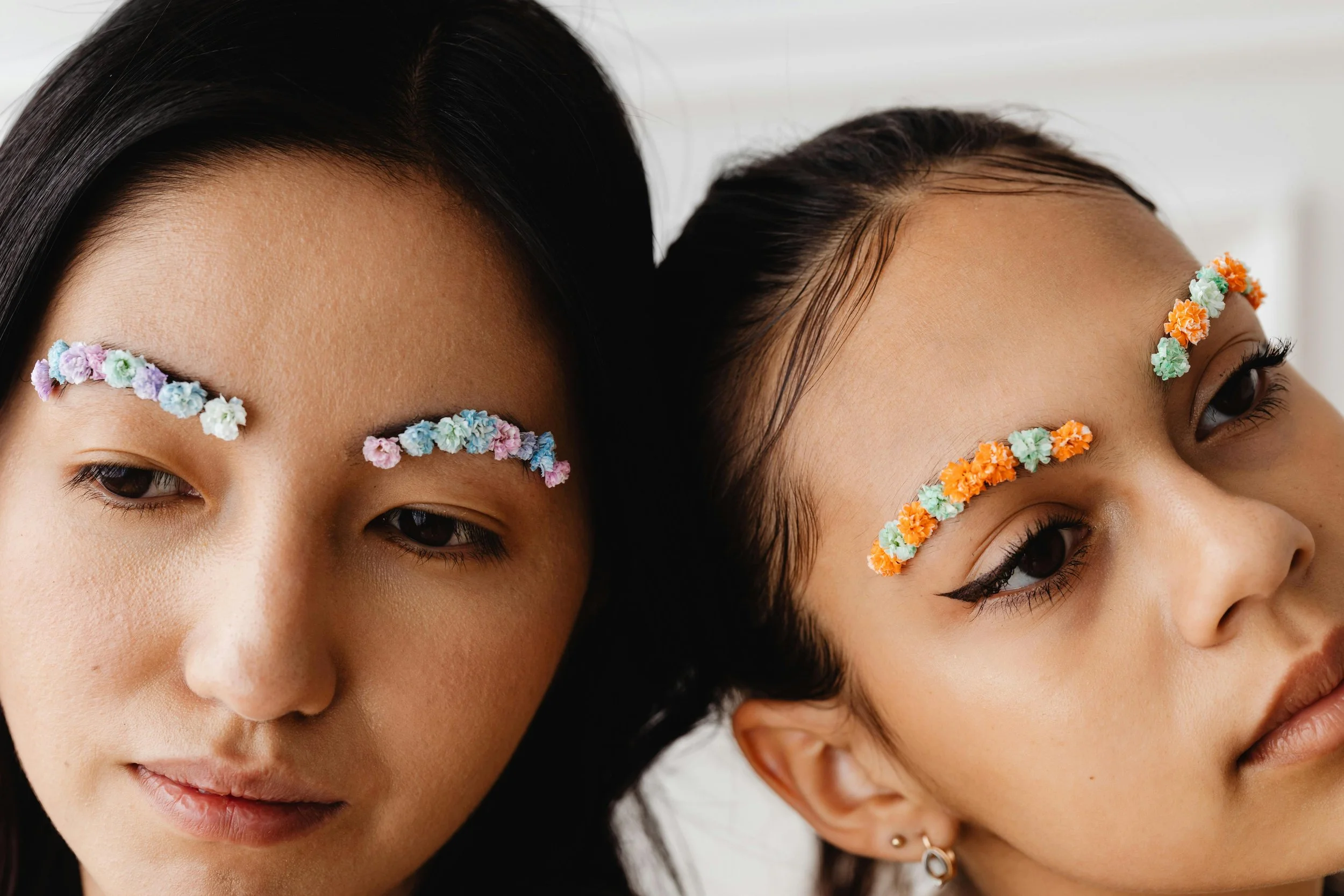Close-up of two women with colorful foam flower face stickers on their eyebrows, lying down with their heads touching. One woman has dark straight hair, and the other has dark wavy hair.