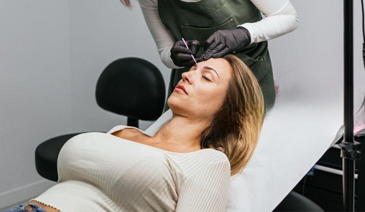 A woman lying down with eyes closed while getting a cosmetic tattooing procedure on her eyebrows by a professional technician.