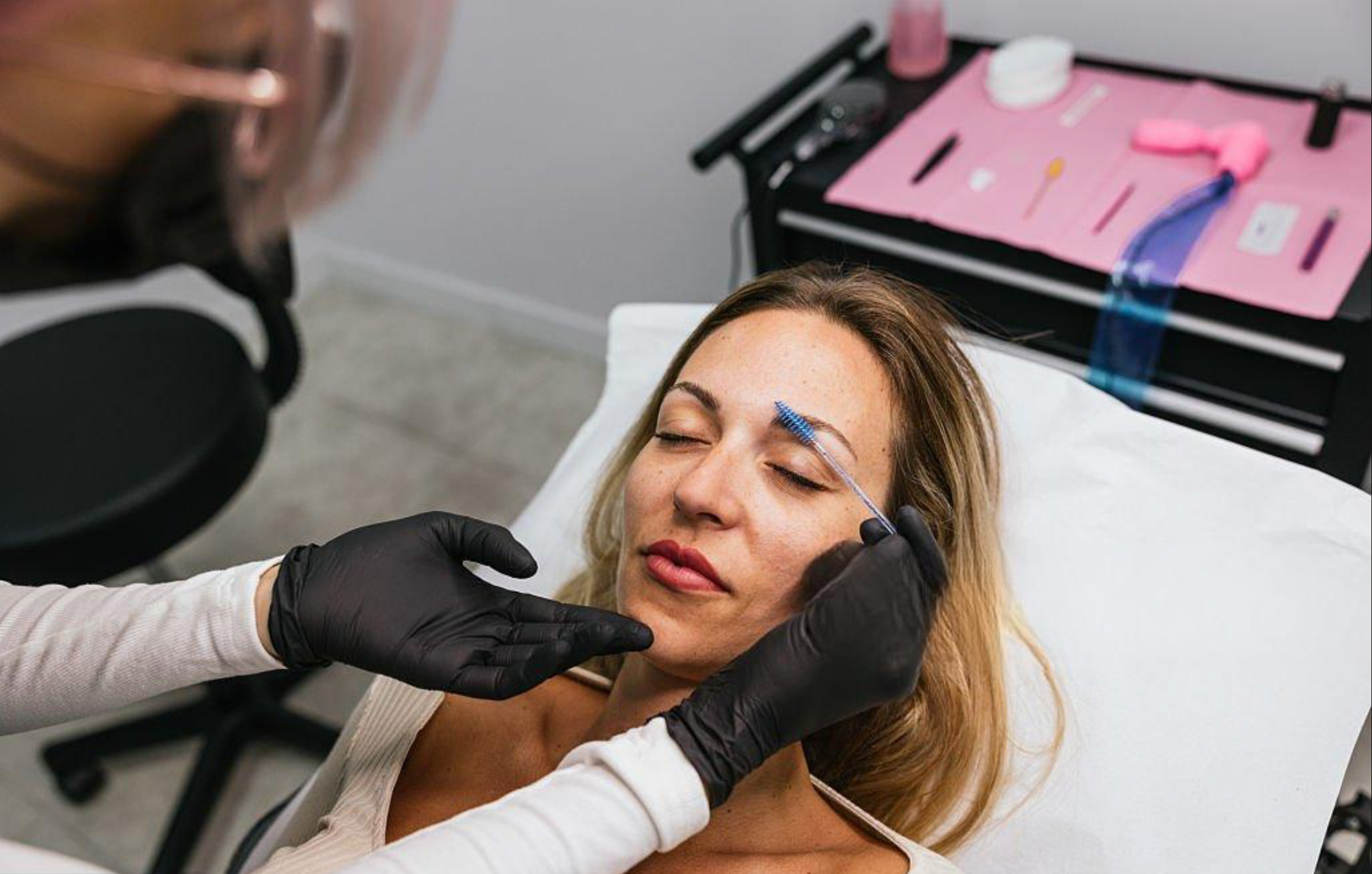 A woman lying down with her eyes closed, receiving cosmetic permanent makeup on her eyebrows from a professional wearing black gloves. A tray with cosmetic tools and supplies is in the background.