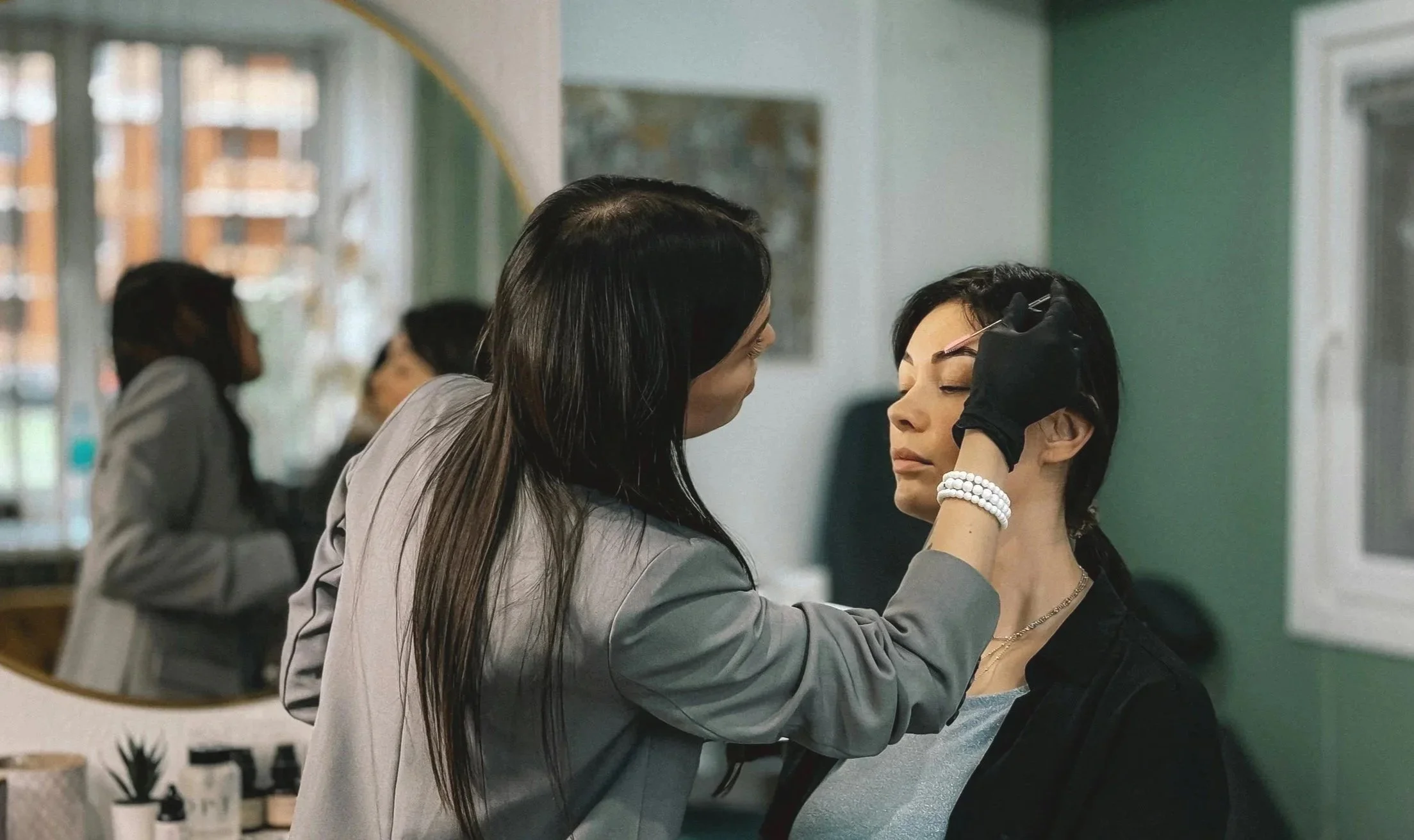 A woman with long dark hair is getting her eyebrows shaped by another woman with dark hair in a salon or beauty studio.