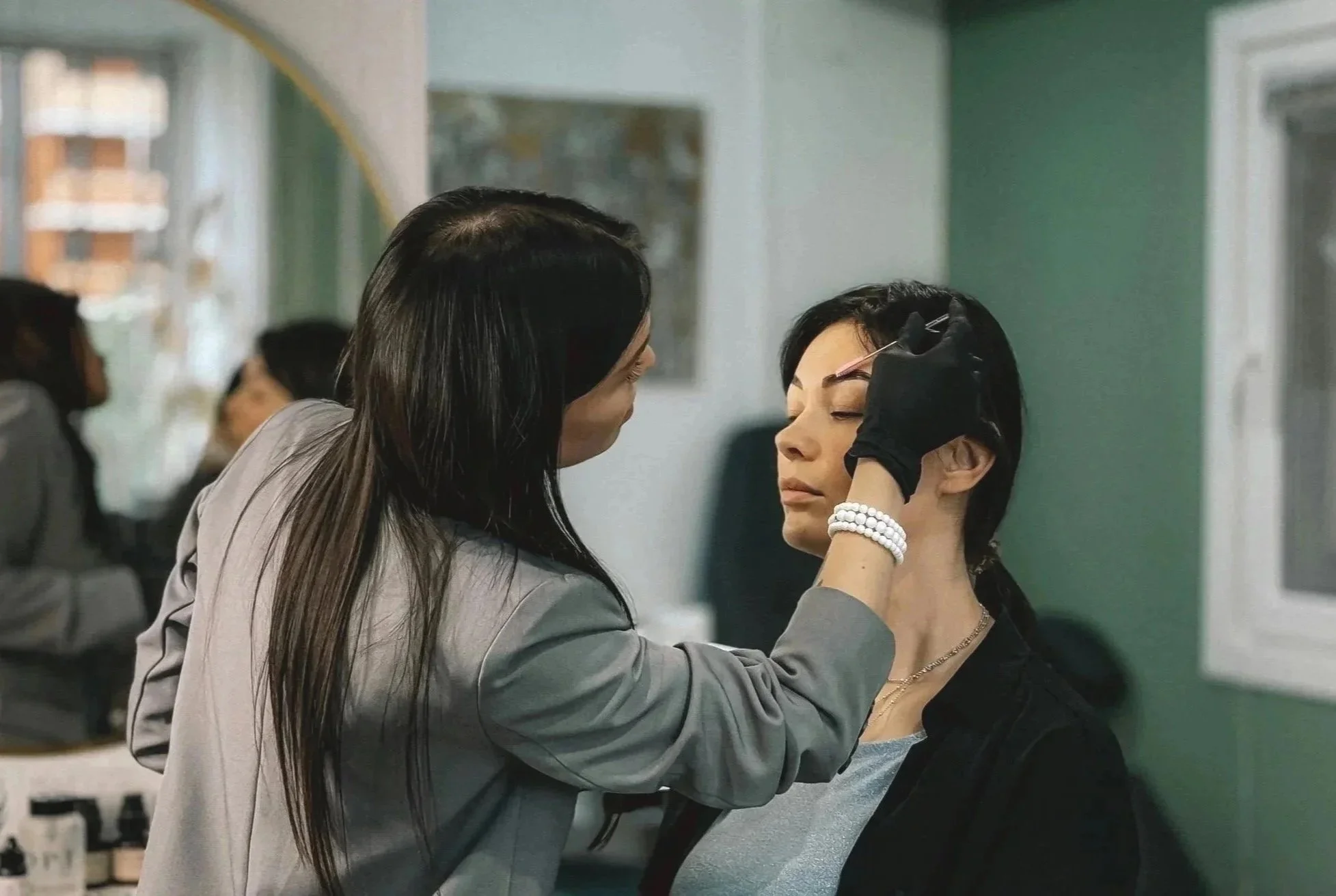 A woman is applying makeup, specifically drawing eyeliner on another woman's eyelid, in a setting resembling a beauty salon or makeup station.