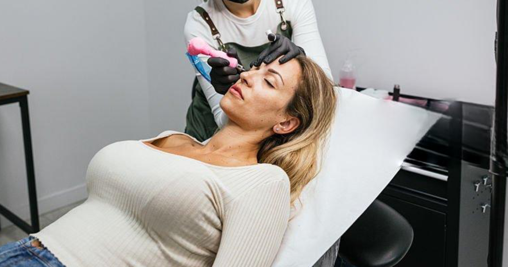 A woman receiving nanoblading on her eyebrows at a tattoo parlor, lying on a tattoo bed with her eyes closed, while a technician uses a tattoo machine.