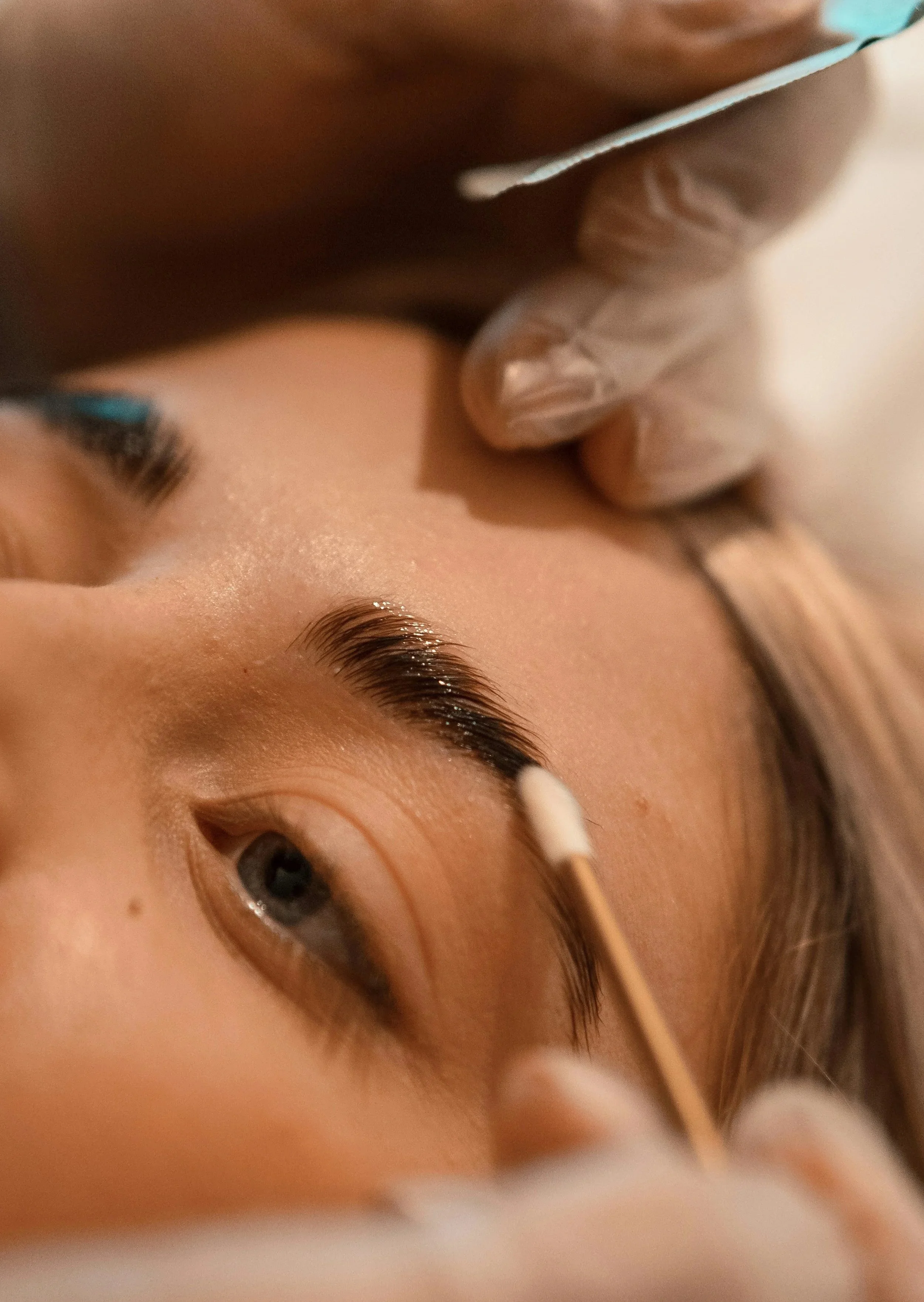 Close-up of a person receiving an eyelash extension procedure, with a technician applying adhesive to the person's eyelashes using a cotton swab.