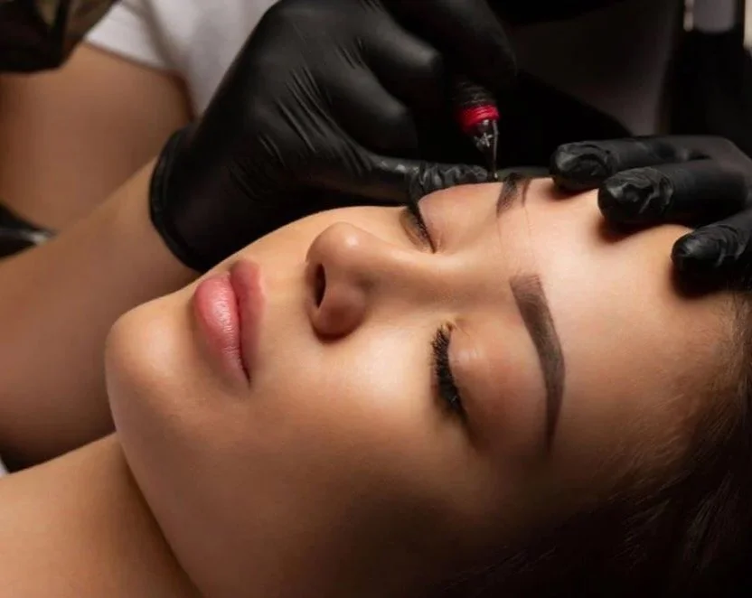 Close-up of woman receiving permanent makeup tattooing on her eyebrows by a technician using a tattoo machine.