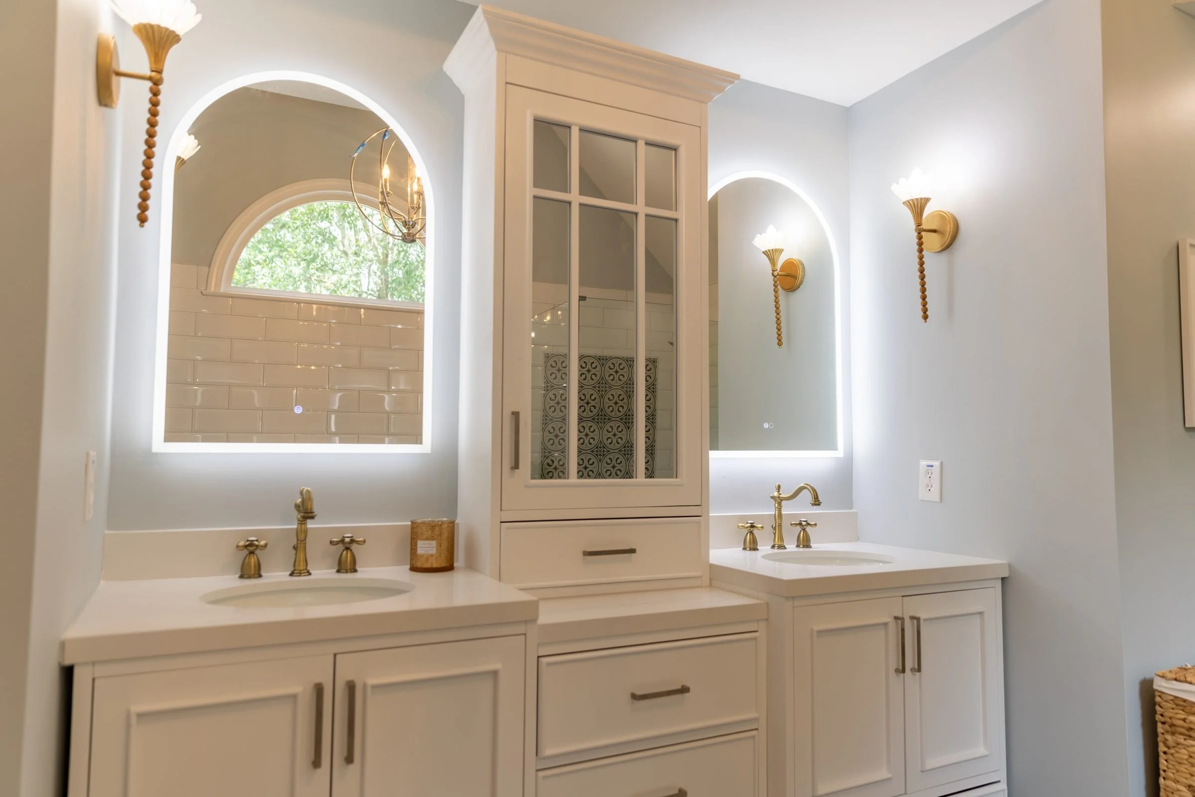 Double vanity with backlit arched mirrors, brass sconces, and glass-front linen cabinet in a Charlotte, NC bathroom remodel