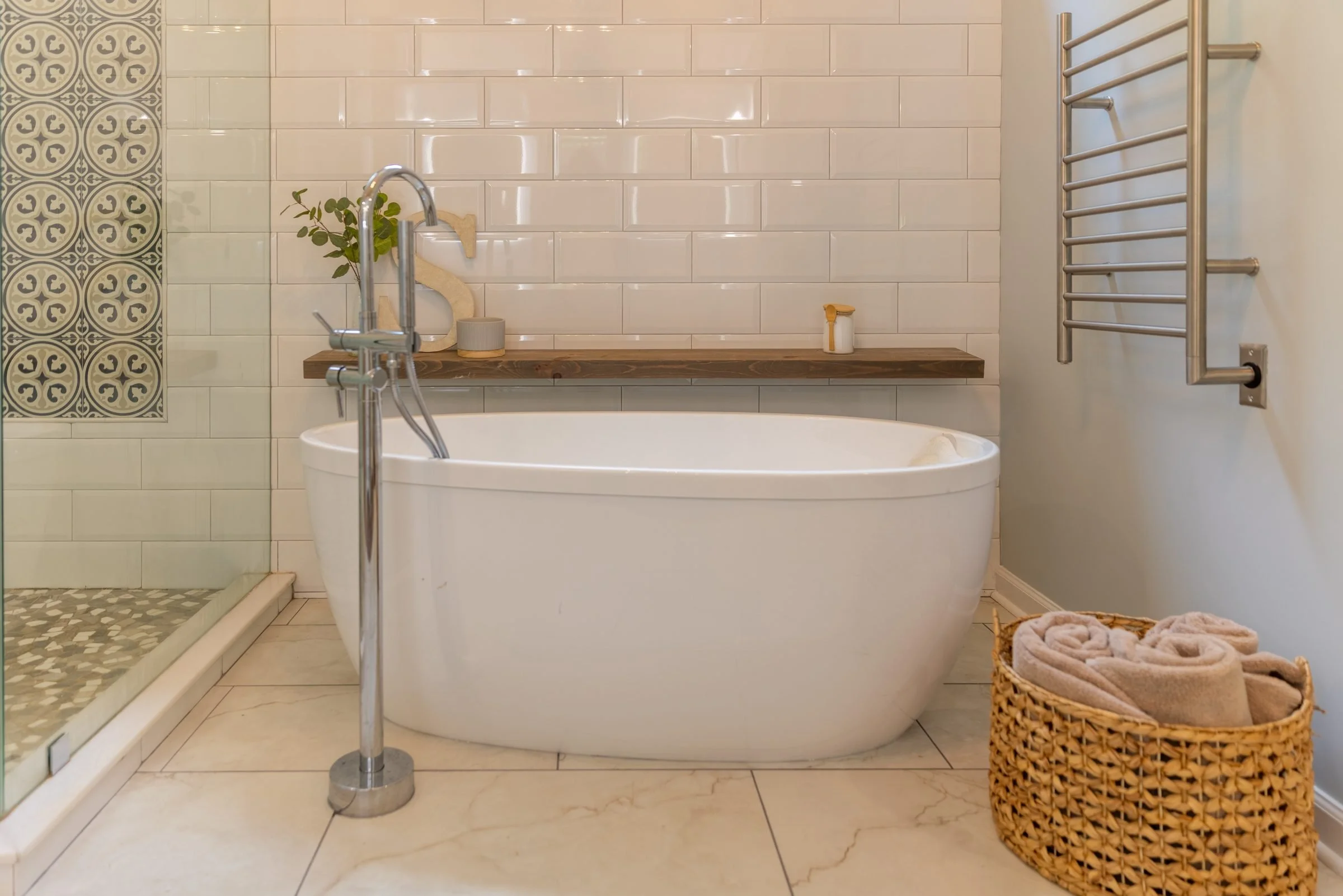 Freestanding soaking tub with floor-mounted faucet and wood shelf against white subway tile in Charlotte, NC