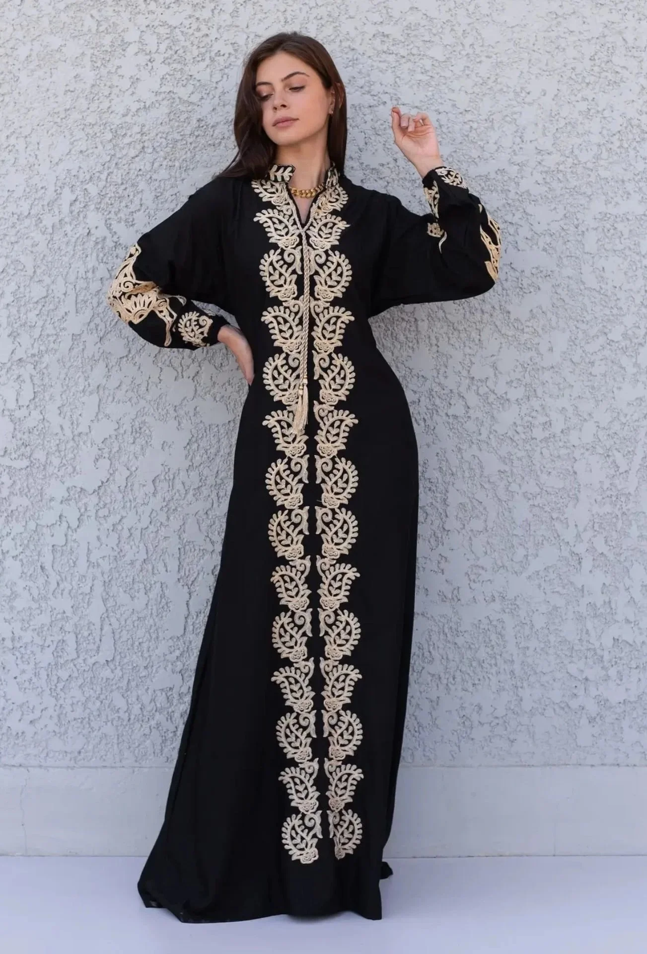 A woman wearing a long black dress with intricate beige embroidery running down the center and on the sleeves, standing against a textured white wall.