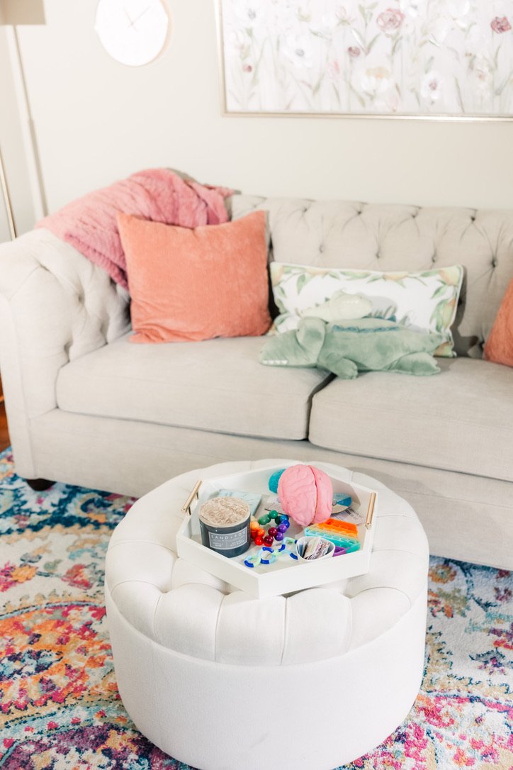 Living room with a beige tufted couch adorned with pastel pillows, a pink throw blanket, and a floral wall art. A white round ottoman with a tray holding colorful toys, a plush brain, and a container. A multicolored floral rug covers the floor.