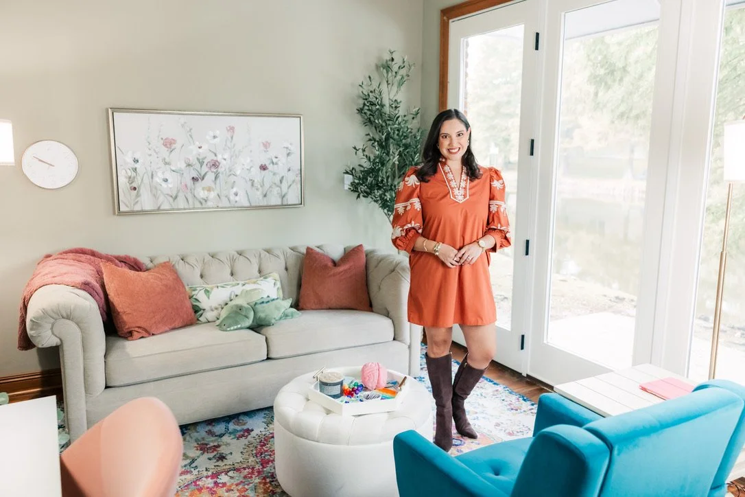 Woman standing in a bright living room near large glass doors, wearing an orange dress and brown knee-high boots, smiling.