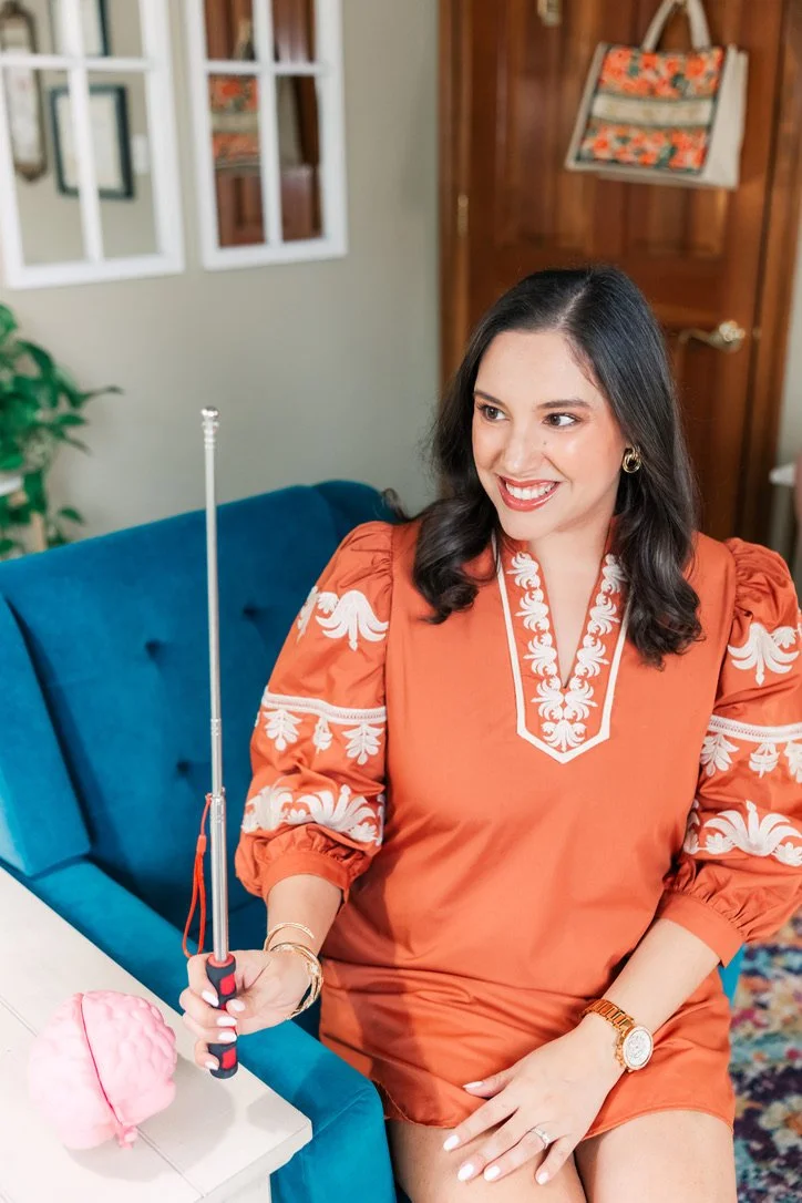 A woman with dark hair, wearing an orange dress with white embroidery, sitting on a teal couch smiling, holding a screwdriver, in a cozy room with wall decor and a wooden door.