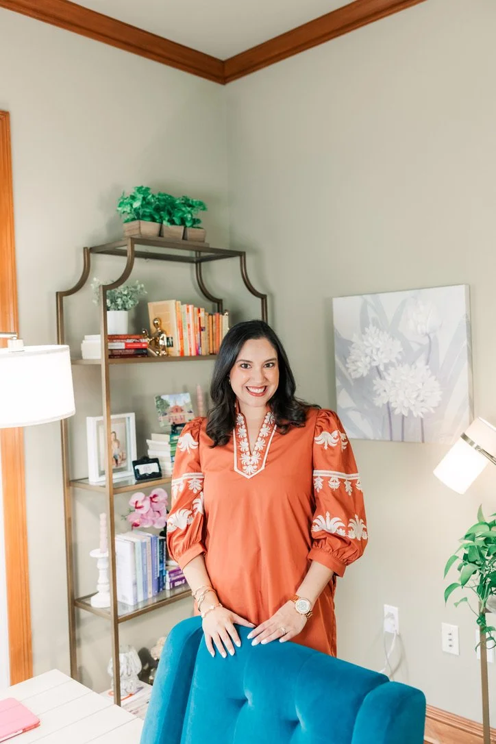 A woman smiling in a room with a bookshelf, a painting, and a green plant. She is wearing an orange top with white embroidery and has dark hair.