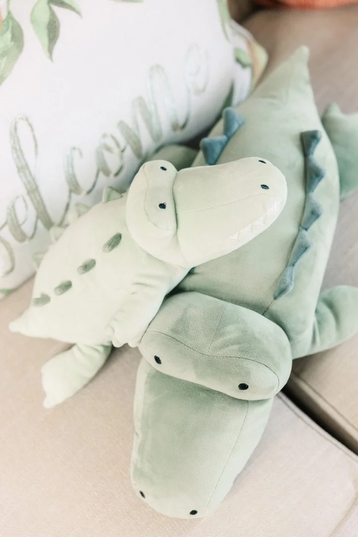 Soft plush crocodile toy resting on a beige surface, with a decorative pillow in the background.