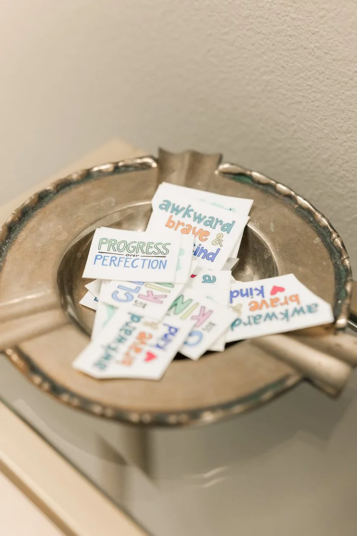 A silver dish with handwritten notes and inspirational quotes such as "awkward brave & kind," "PROGRESS over PERFECTION," and "flawed and loved," resting on a white surface against a beige wall.