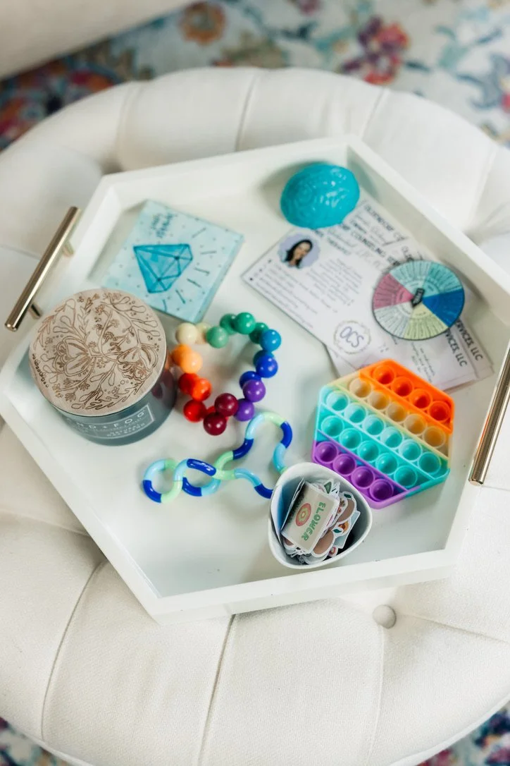 A white tray filled with colorful objects including a rainbow beaded bracelet, rainbow-colored fidget toy, card with blue diamond, blue decorative object, personalized business card, pie chart, and a small container of stickers. The tray is placed on