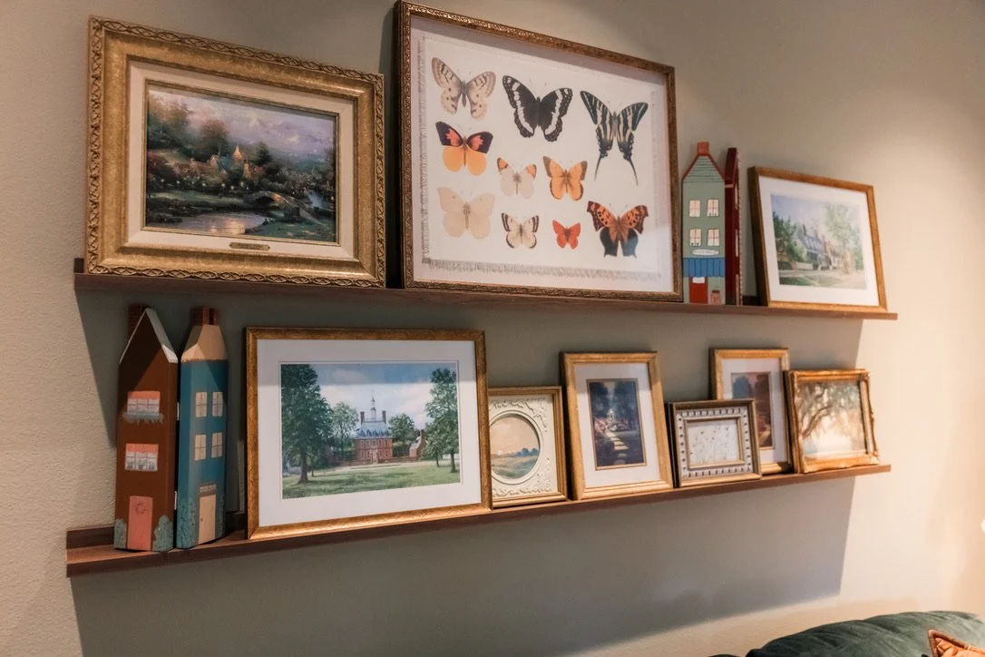Two wooden shelves holding framed pictures, artwork including butterfly prints, decorative houses, and landscape paintings, arranged on a beige wall.